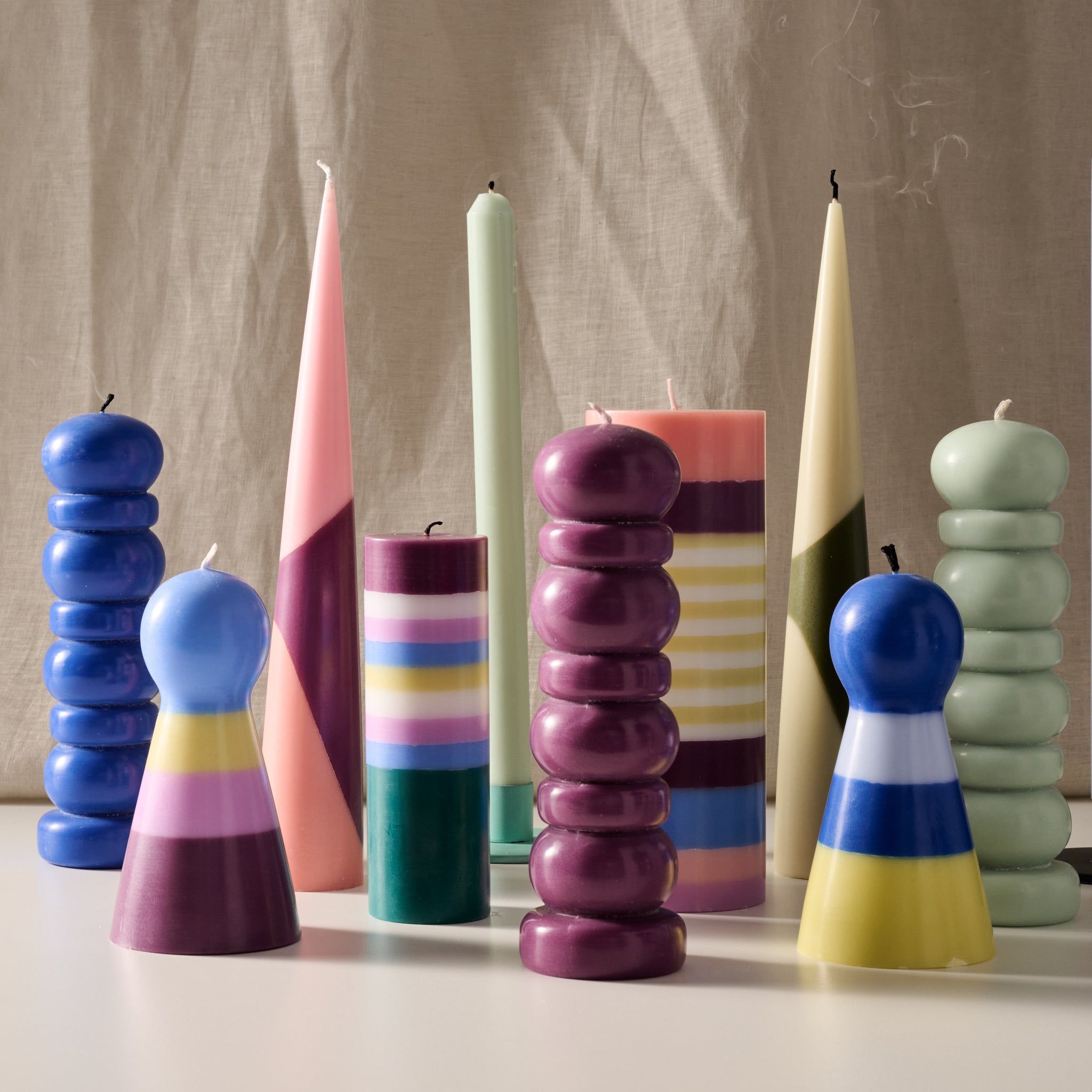 A group of colorful sculptural taper candles and pillar candles arranged on a white surface against a beige fabric backdrop. The candles feature bold geometric and striped patterns in shades of blue, pink, purple, green, yellow, and white.
