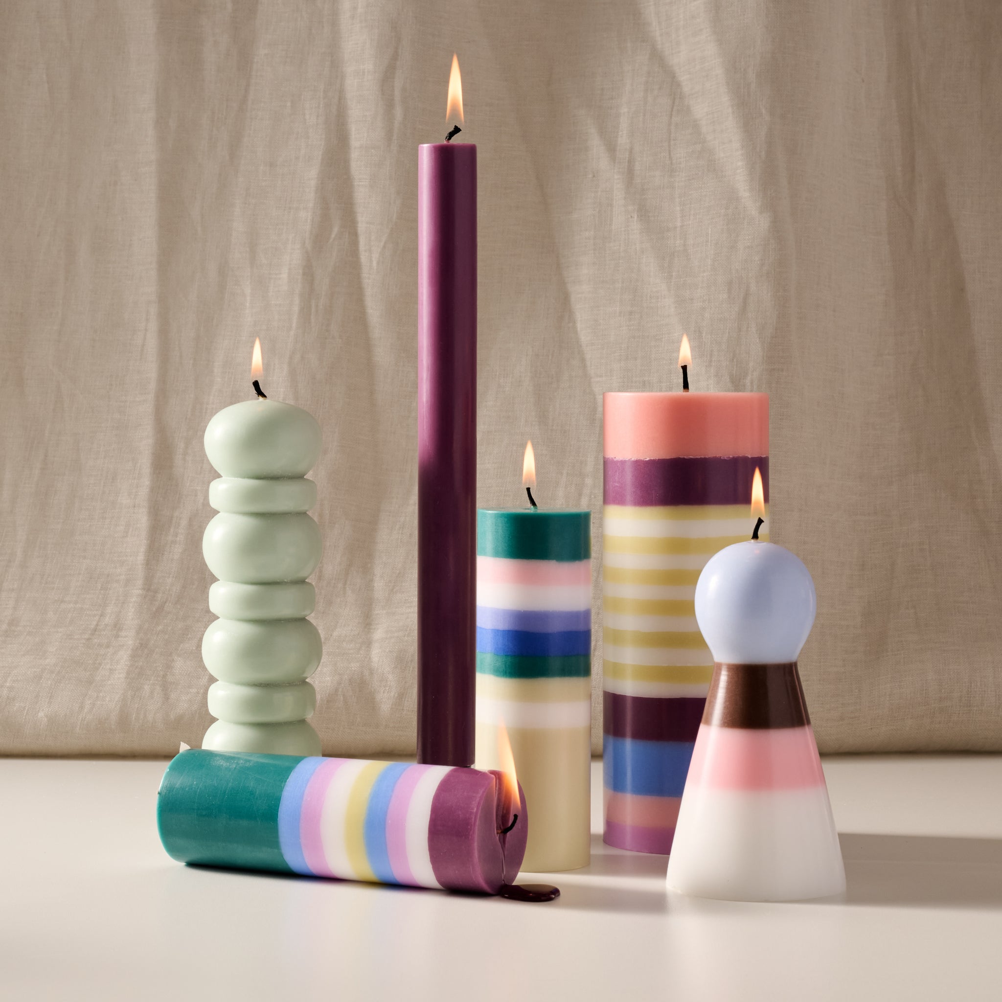 A group of colorful sculptural candles arranged on a white surface against a beige fabric backdrop. The candles include stacked round tiers in pale mint, a single tall purple candle, striped candles in teal/blue/white, cream/yellow, and pink/blue, and a cone-shaped candle with a light blue top.