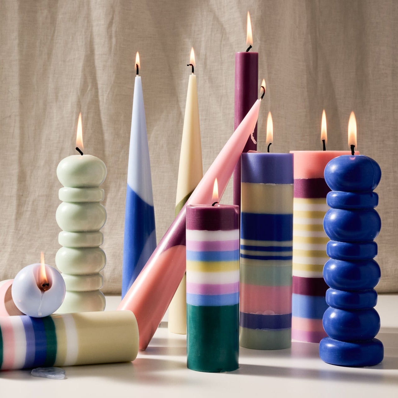 A group of colorful decorative candles, some lit with visible flames, arranged on a light surface against a beige fabric backdrop. The candles have varied shapes, including stacked discs, rounded towers, and striped cylinders, in pastel and saturated colors such as blue, pink, purple, green, yellow, and white.