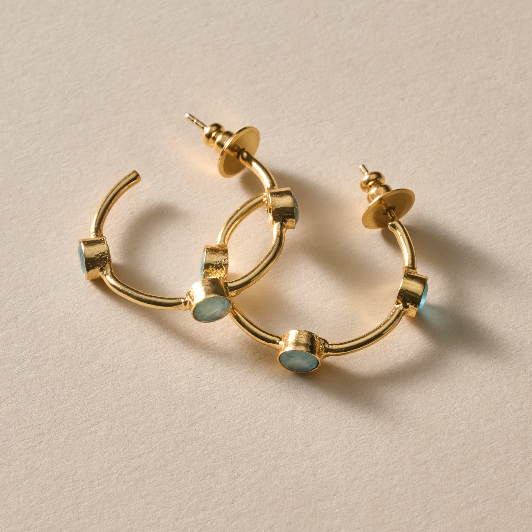 Gold hoop earrings with small oval light blue inlay accents, photographed on a warm beige textured background.