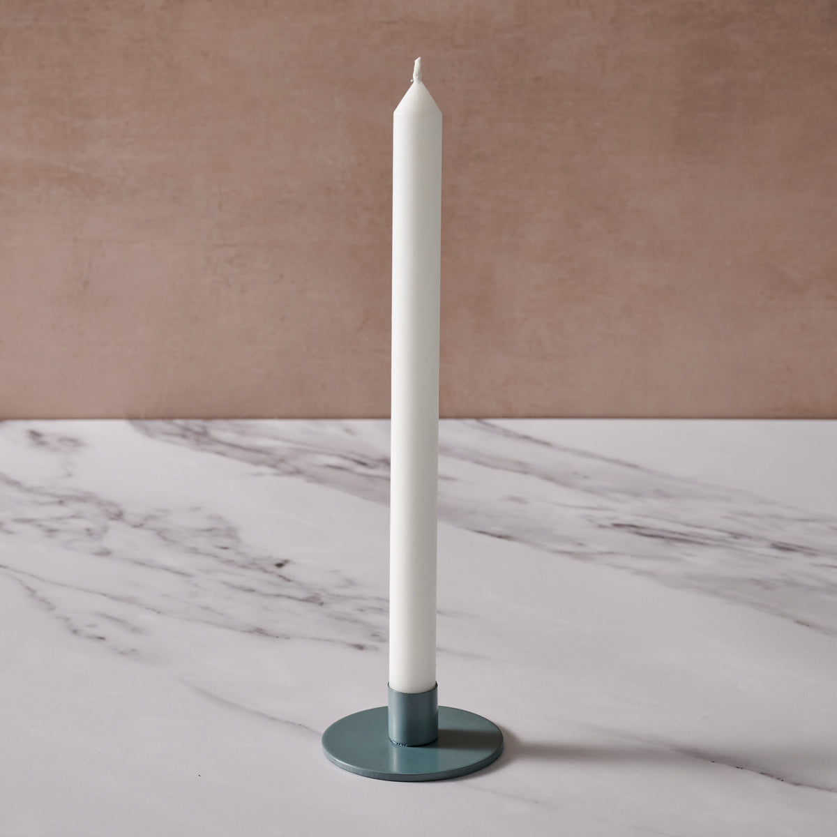 A tall white taper candle in a round blue/gray metal holder, set on a white marble surface against a textured beige-brown background.