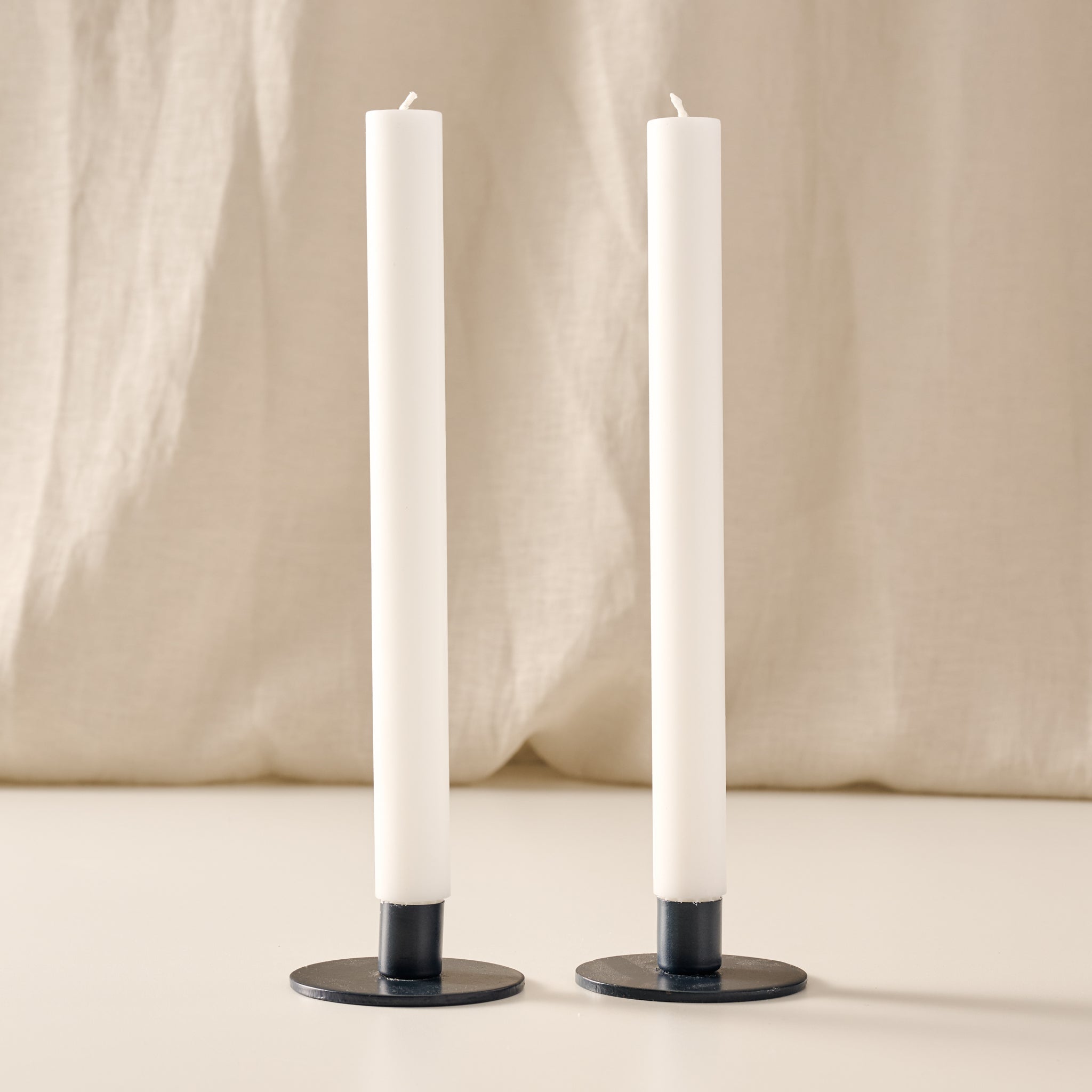 Two tall natural white taper candles in round blue metal holders, standing on a light surface against a soft beige fabric backdrop.