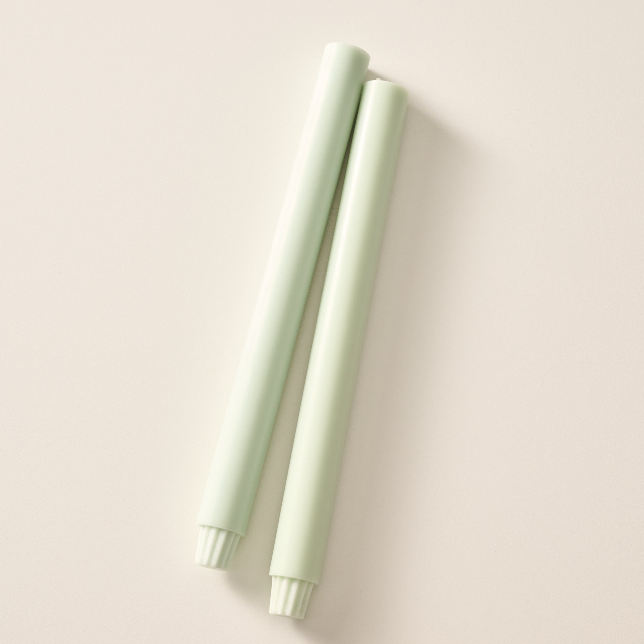 Two sage green colored pillar candles with ribbed bases, laying flat on a white background.