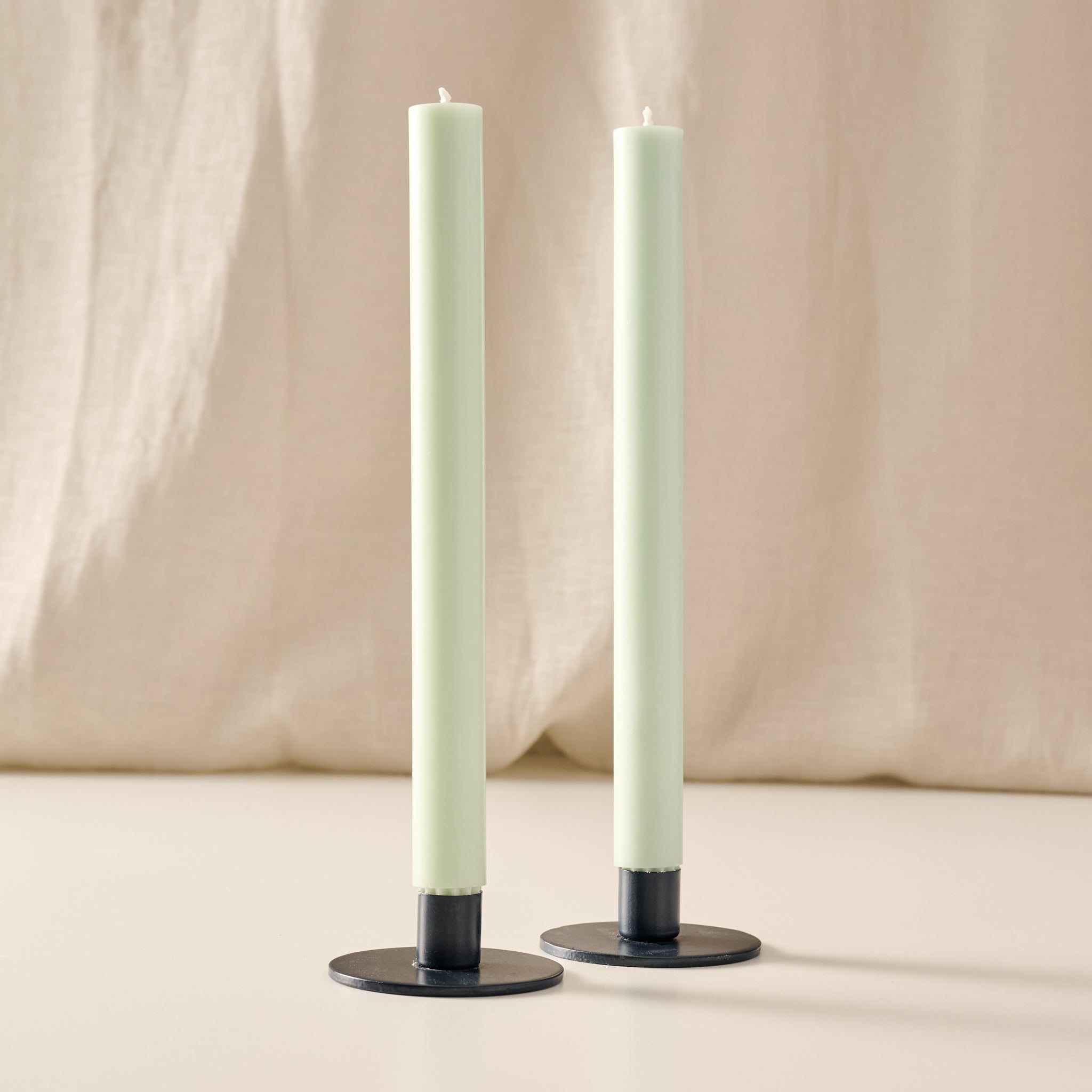 Two tall, slim pillar candles in a sage green color, standing in blue metal candle holders with round bases against a soft beige fabric backdrop.