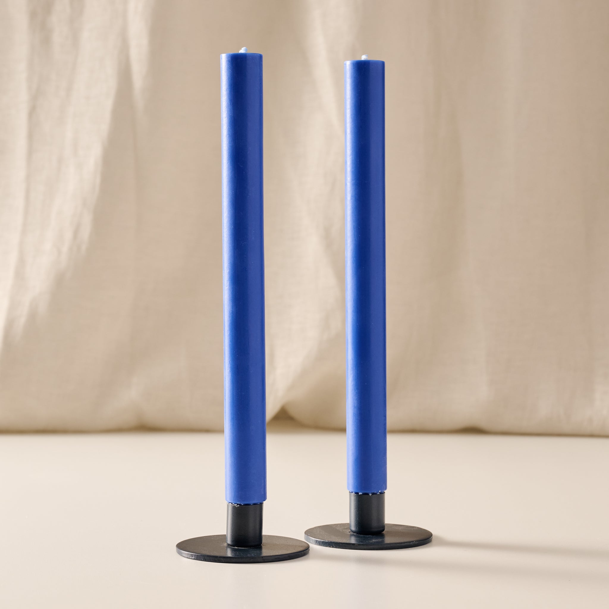Two tall cobalt blue taper candles in blue metal holders, standing upright against a soft beige fabric backdrop.