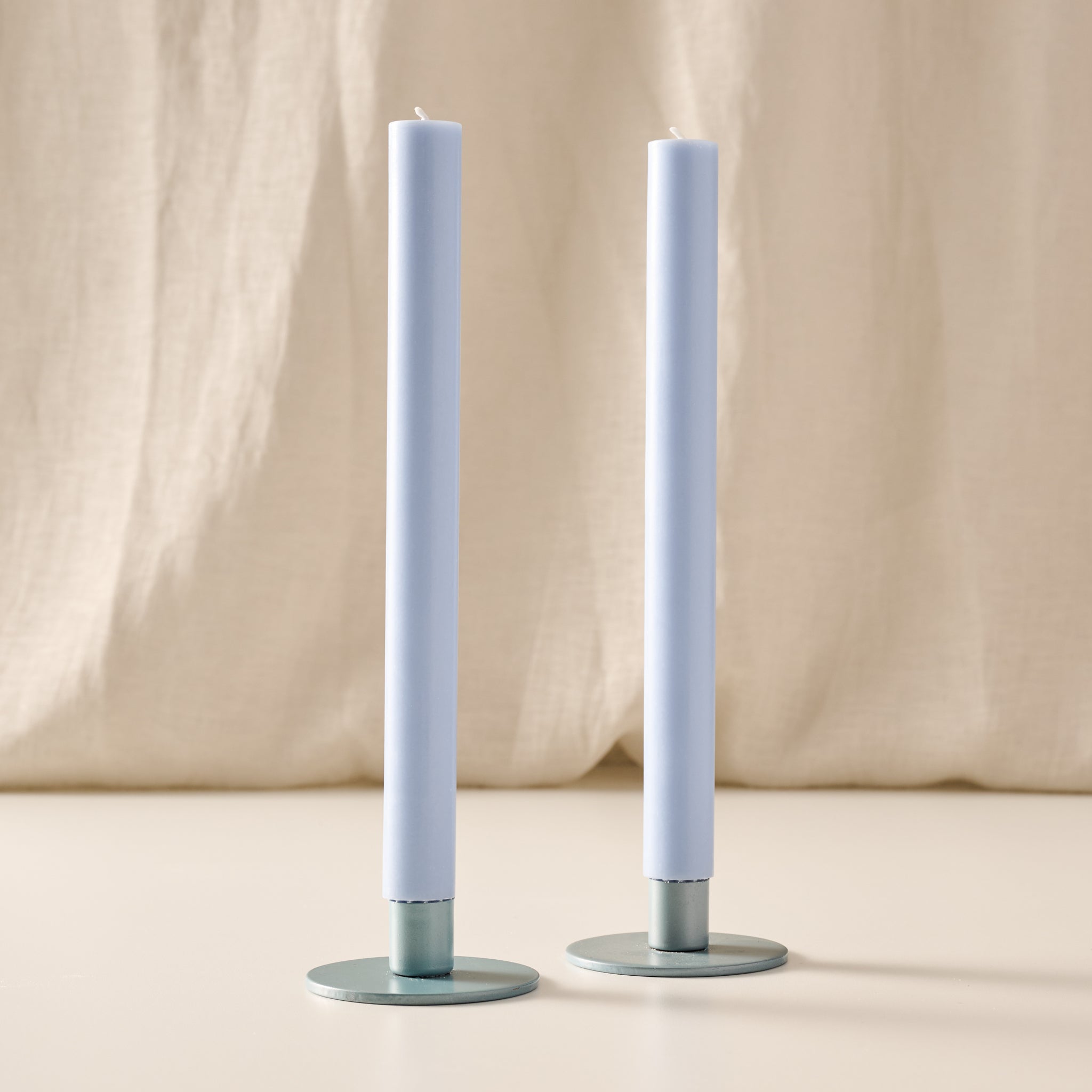 Two tall mint taper candles in round blue/gray holders, standing on a light beige surface with a draped beige fabric backdrop.