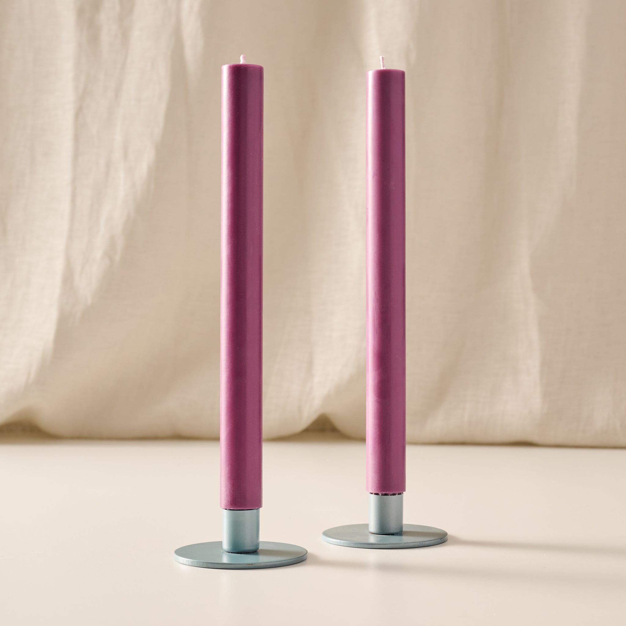 Two slim blackberry dinner candles in round blue/gray metal holders, standing on a light surface against a draped cream fabric backdrop.