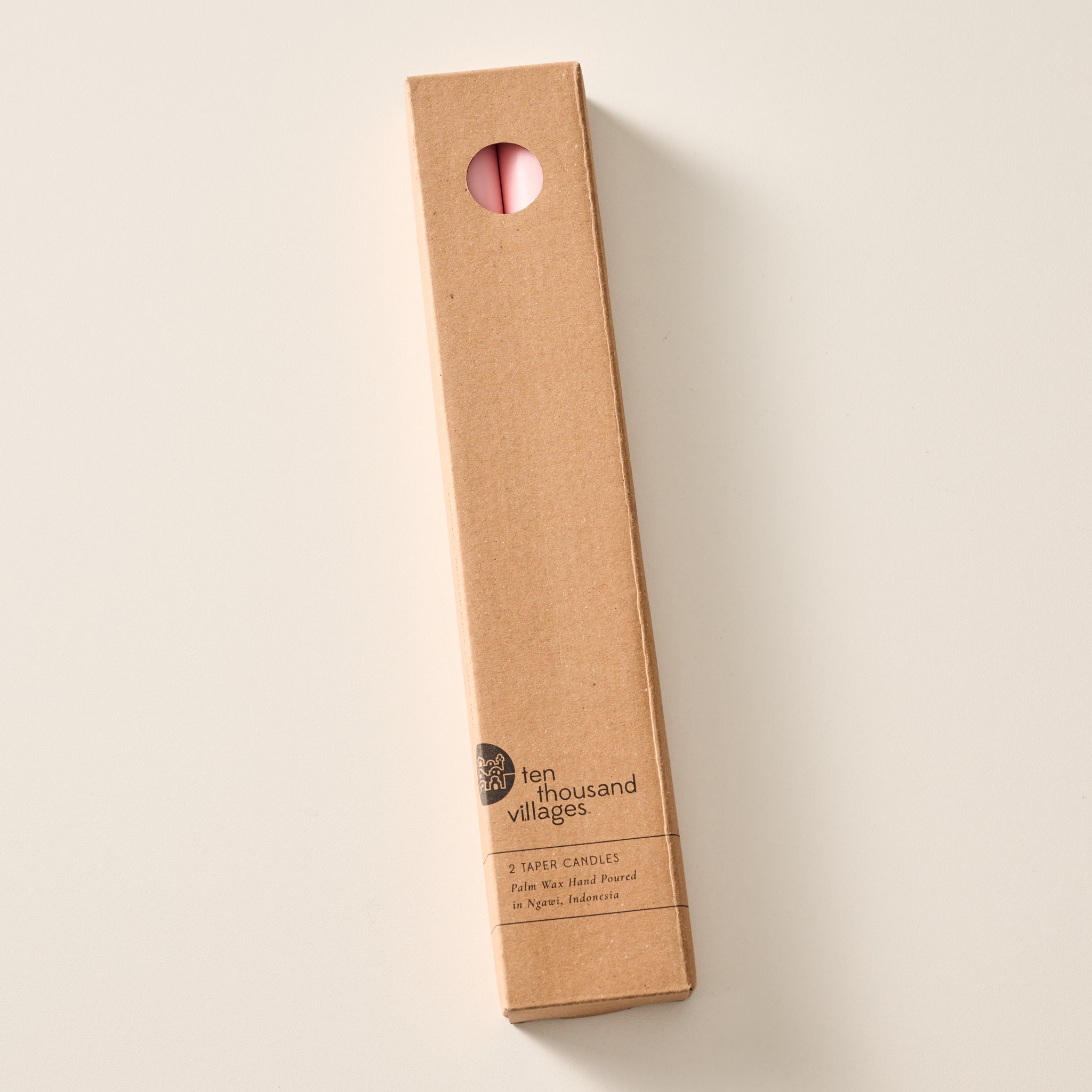 A tall, narrow cardboard box with a circular window display cutout at the top showing two ginger pink candles inside, photographed on a clean off-white background. The box has printed text near the bottom including the brand name 'Ten Thousand Villages'.