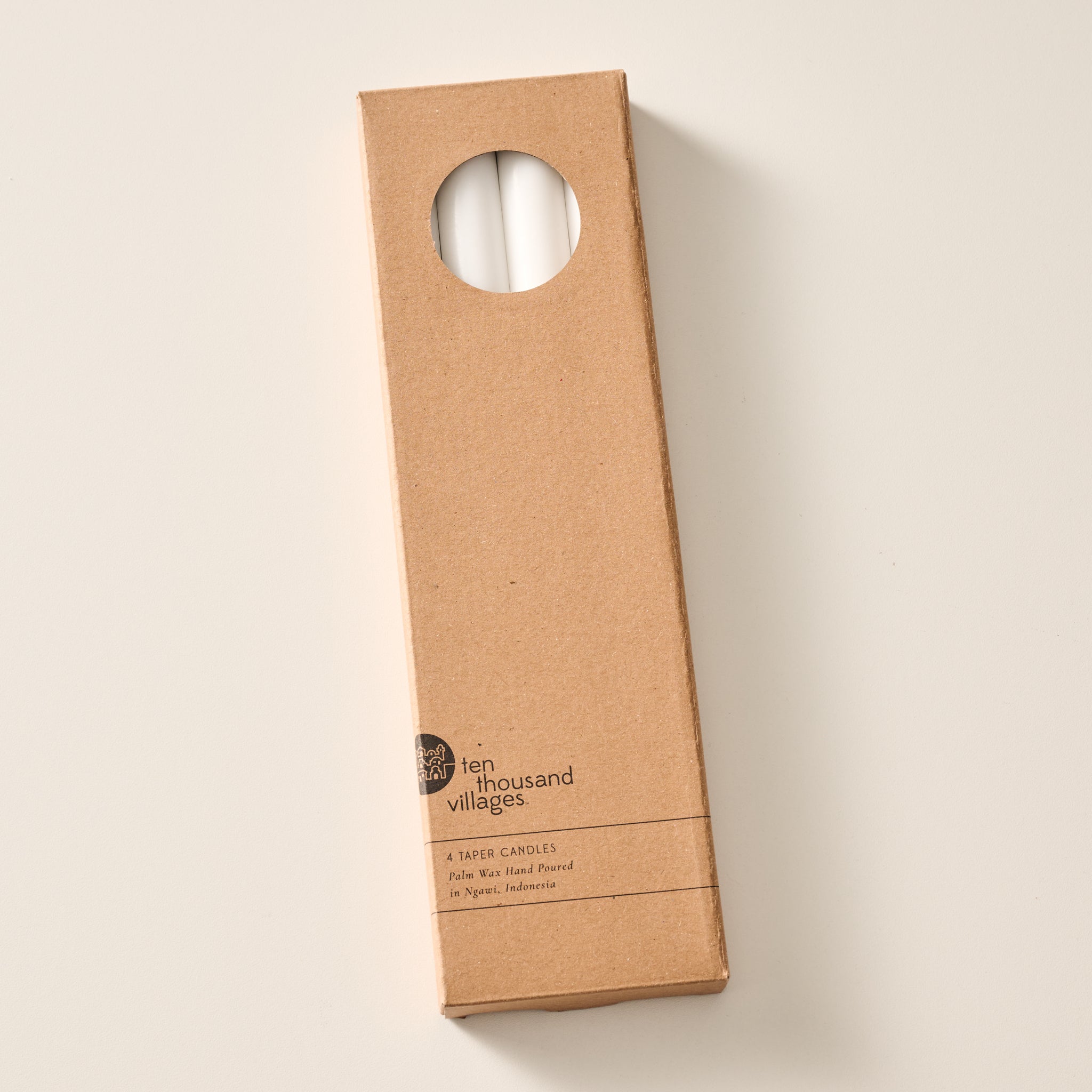 A tall, narrow cardboard box with a circular window display cutout at the top showing two natural white candles inside, photographed on a clean off-white background. The box has printed text near the bottom including the brand name 'Ten Thousand Villages'.