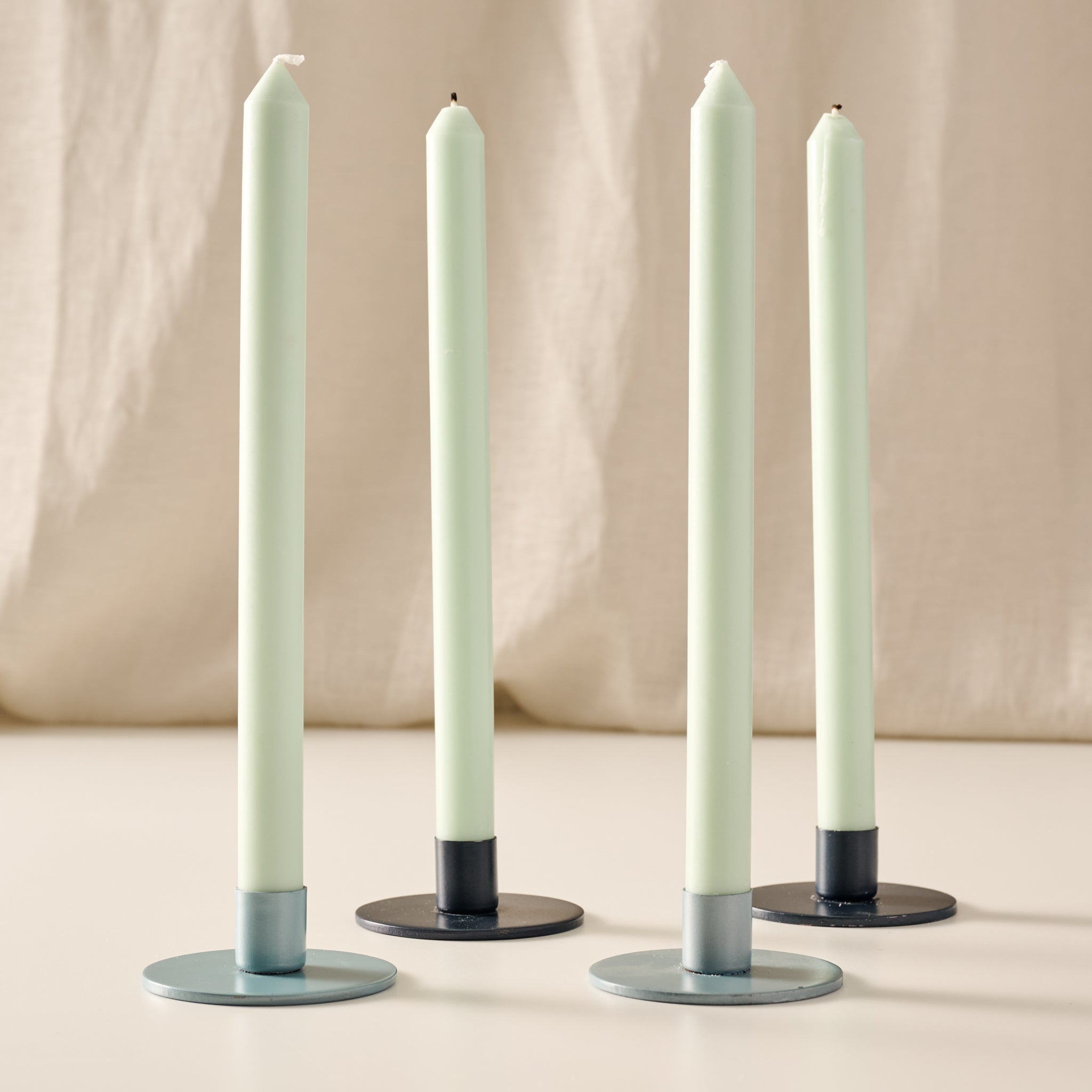 Four taper candles in a sage green color, each resting in a candle holder in the colors of blue/gray and blue, set against a soft beige fabric backdrop.