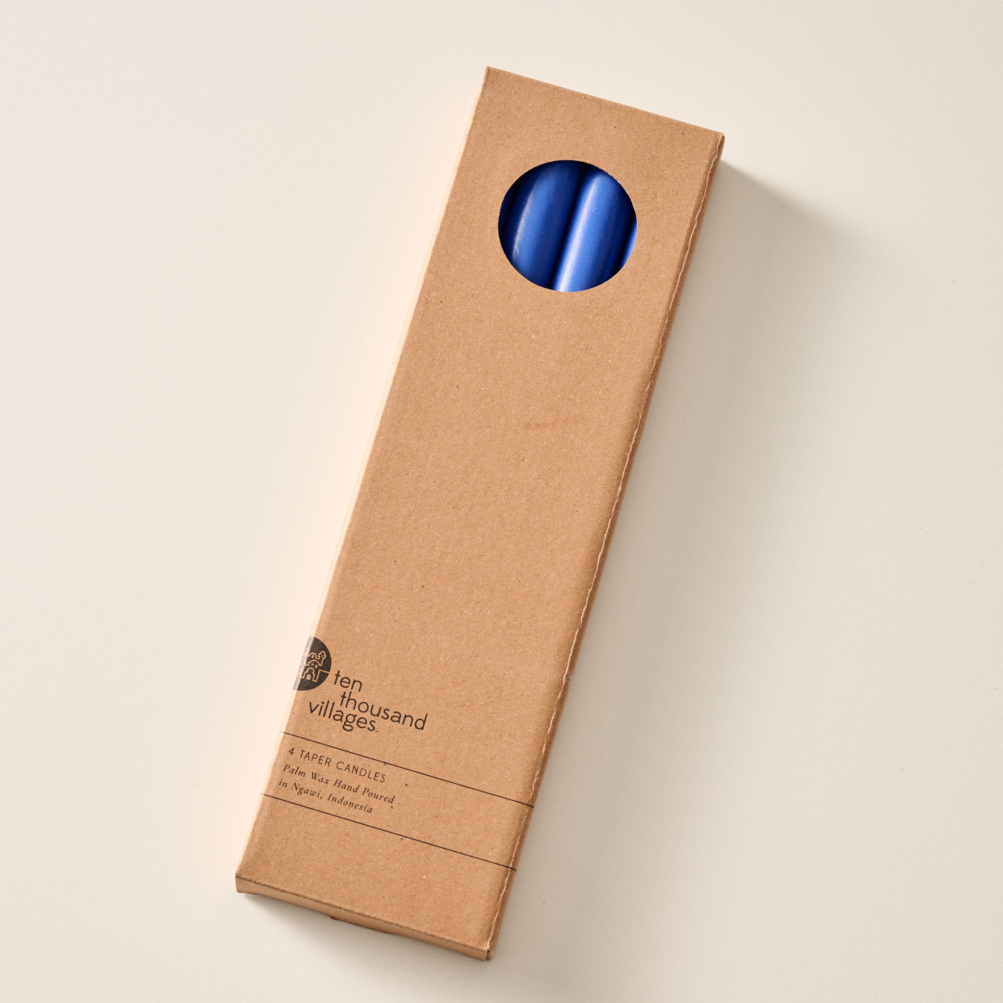 A tall, narrow cardboard box with a circular window display cutout at the top showing two cobalt blue candles inside, photographed on a clean off-white background. The box has printed text near the bottom including the brand name 'Ten Thousand Villages'.
