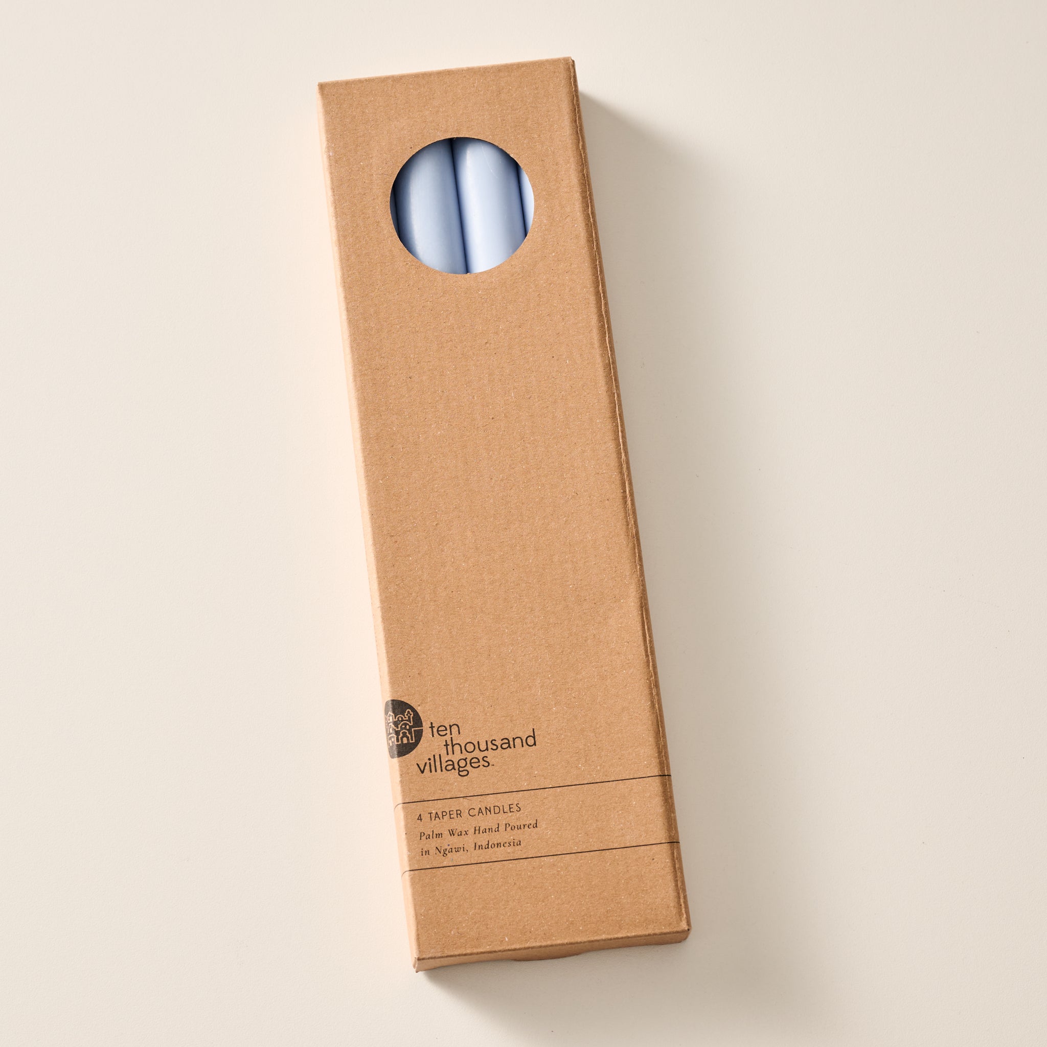 A tall, narrow cardboard box with a circular window display cutout at the top showing two mint colored candles inside, photographed on a clean off-white background. The box has printed text near the bottom including the brand name 'Ten Thousand Villages'.