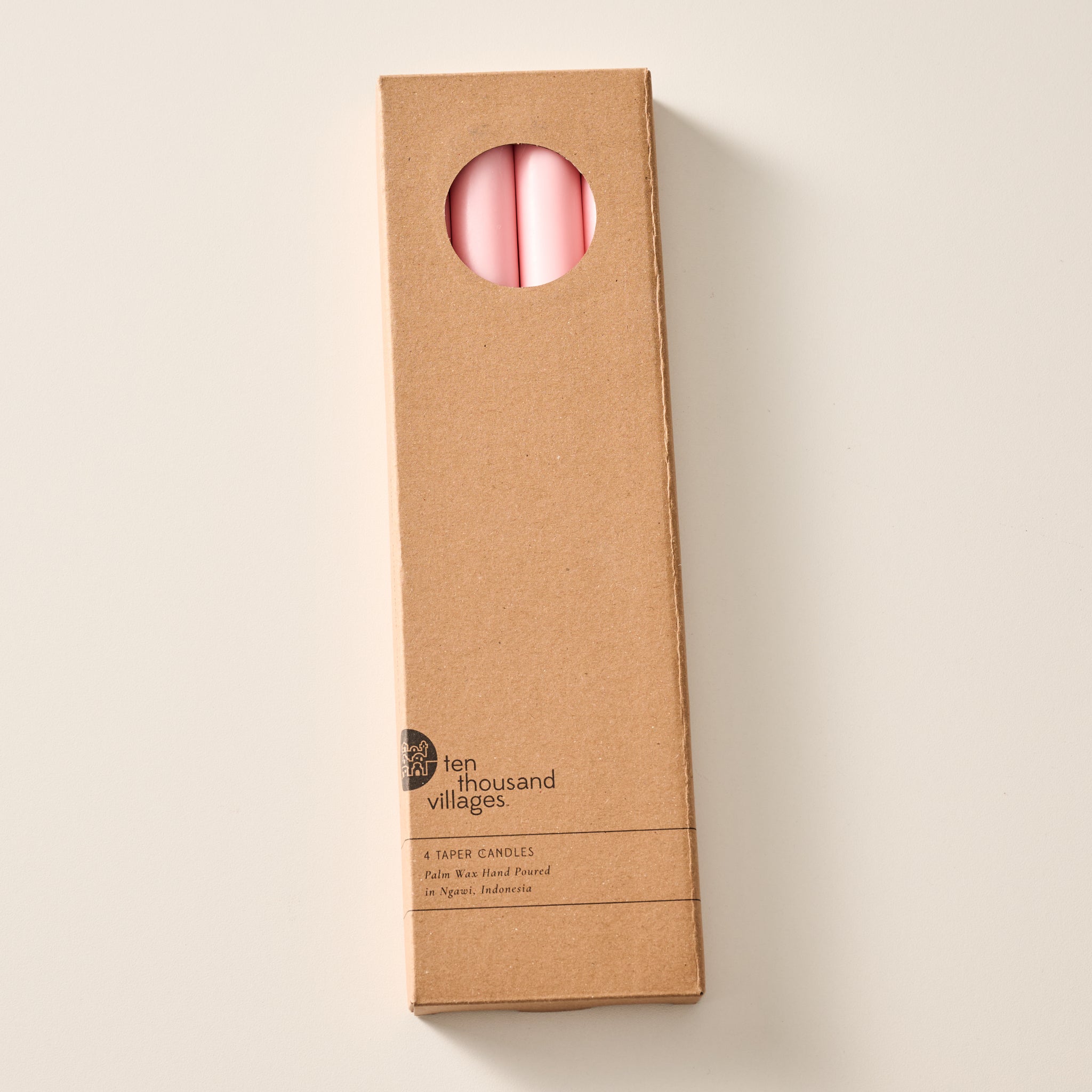A tall, narrow cardboard box with a circular window display cutout at the top showing two ginger colored candles inside, photographed on a clean off-white background. The box has printed text near the bottom including the brand name 'Ten Thousand Villages'.