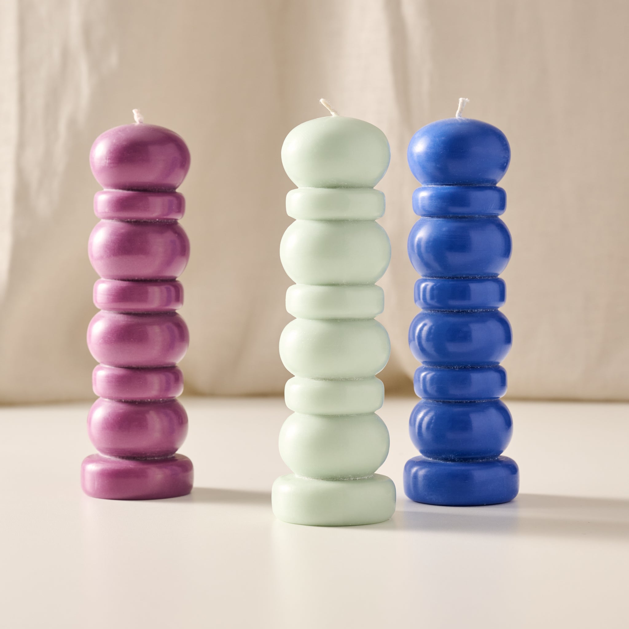 Three stacked, ribbed pillar candles in purple, mint green, and blue, standing on a light surface against a soft beige fabric backdrop.