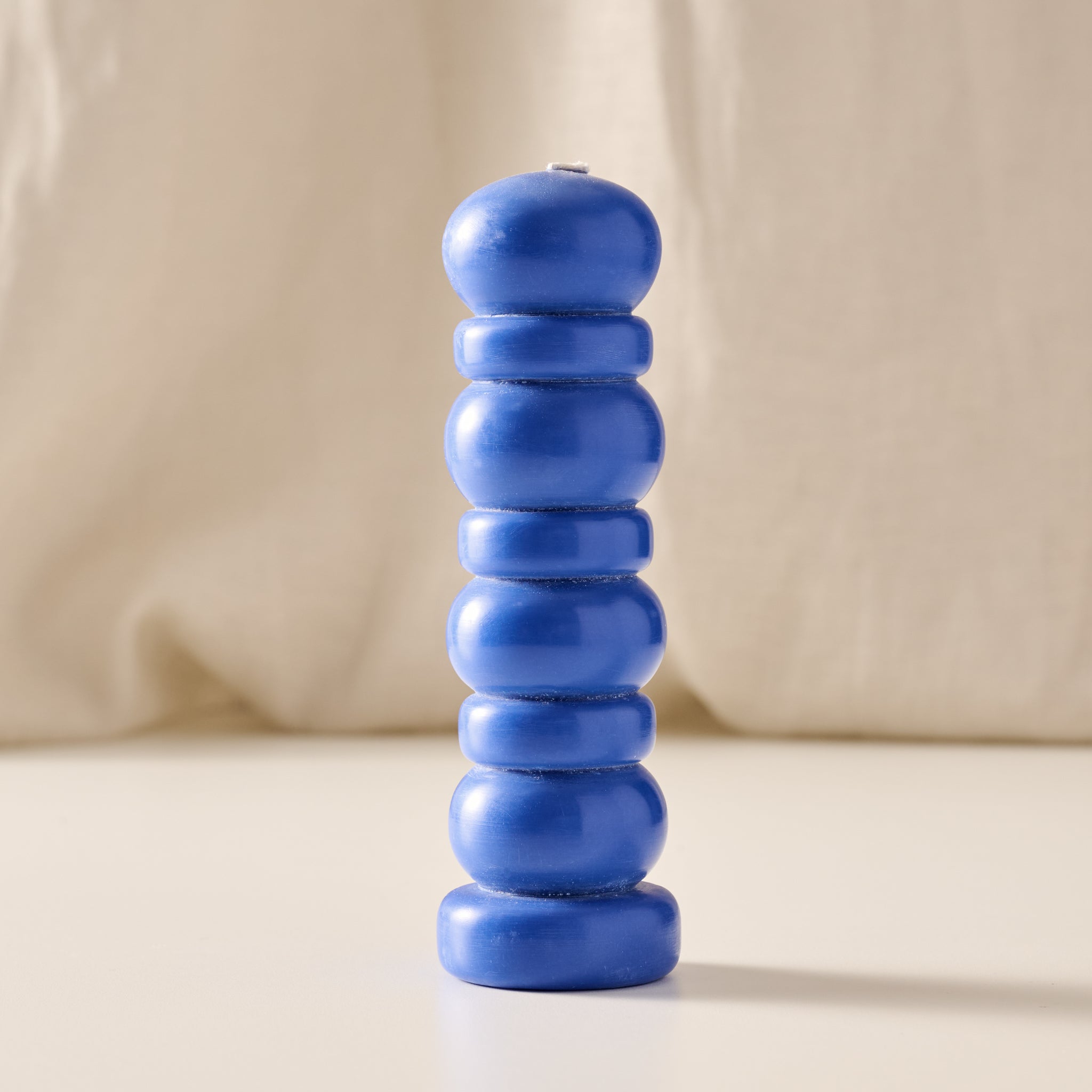 A blue segmented pillar candle with a rounded, stacked design, standing upright on a light beige surface against a soft beige fabric backdrop.