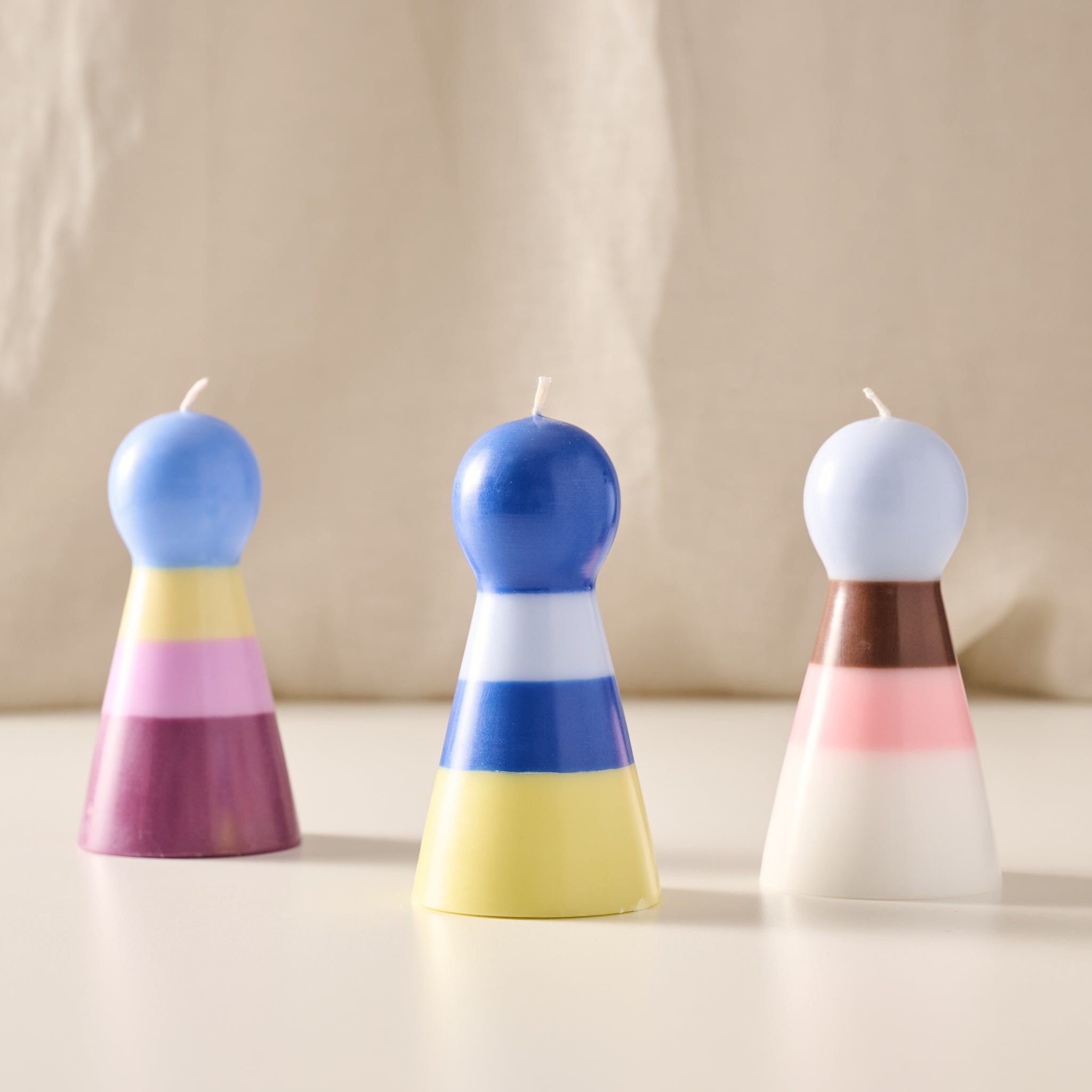 Three pawn-shaped candles with rounded tops are arranged on a light surface against a soft beige fabric backdrop. The center candle is blue, white, and yellow, while the left and right candles have different color combinations including blue, pink, purple, brown, and white.