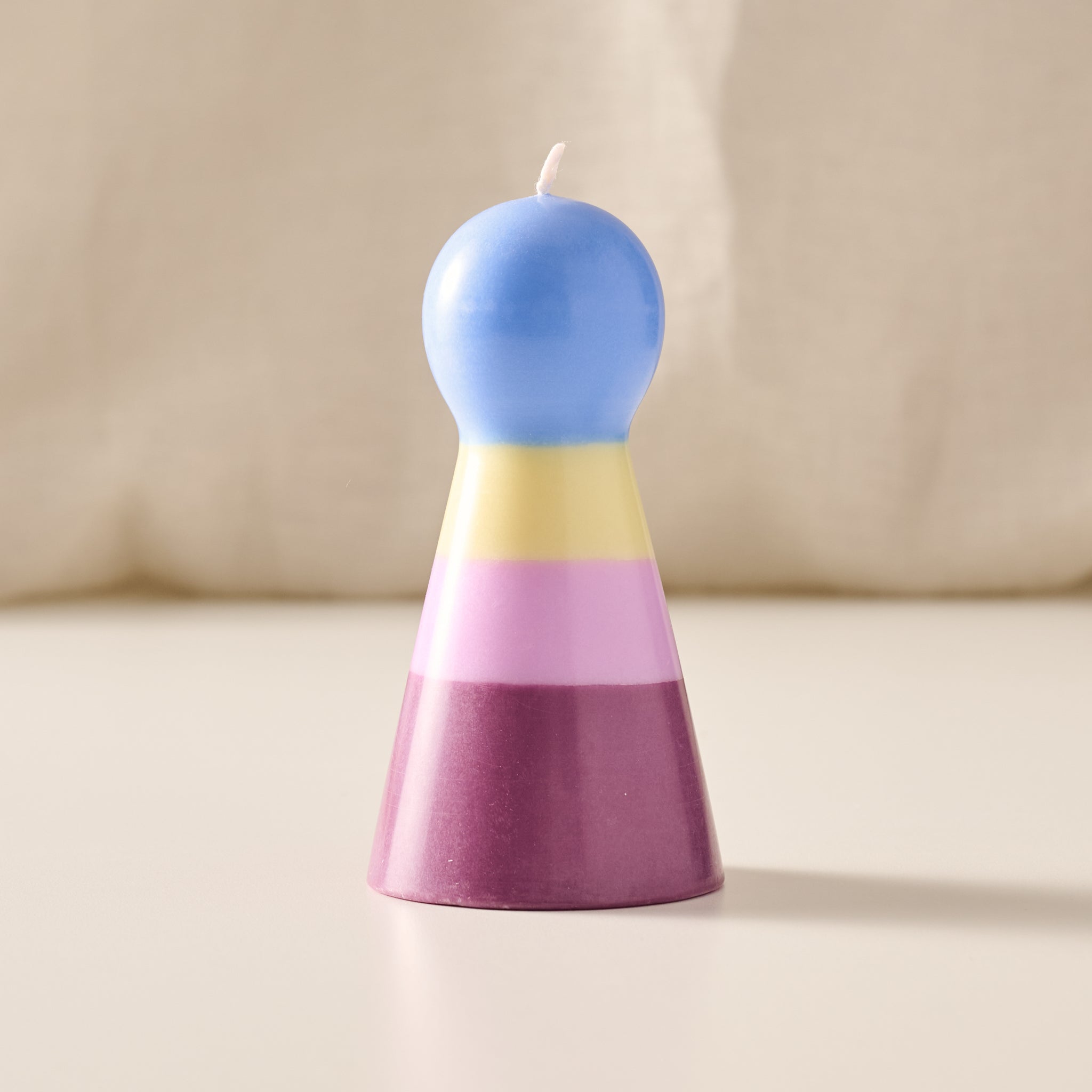 A pawn shaped candle with a rounded blue top and a tapered base in horizontal pastel stripes of yellow, pink, and purple, standing on a light beige surface against a soft cream background.