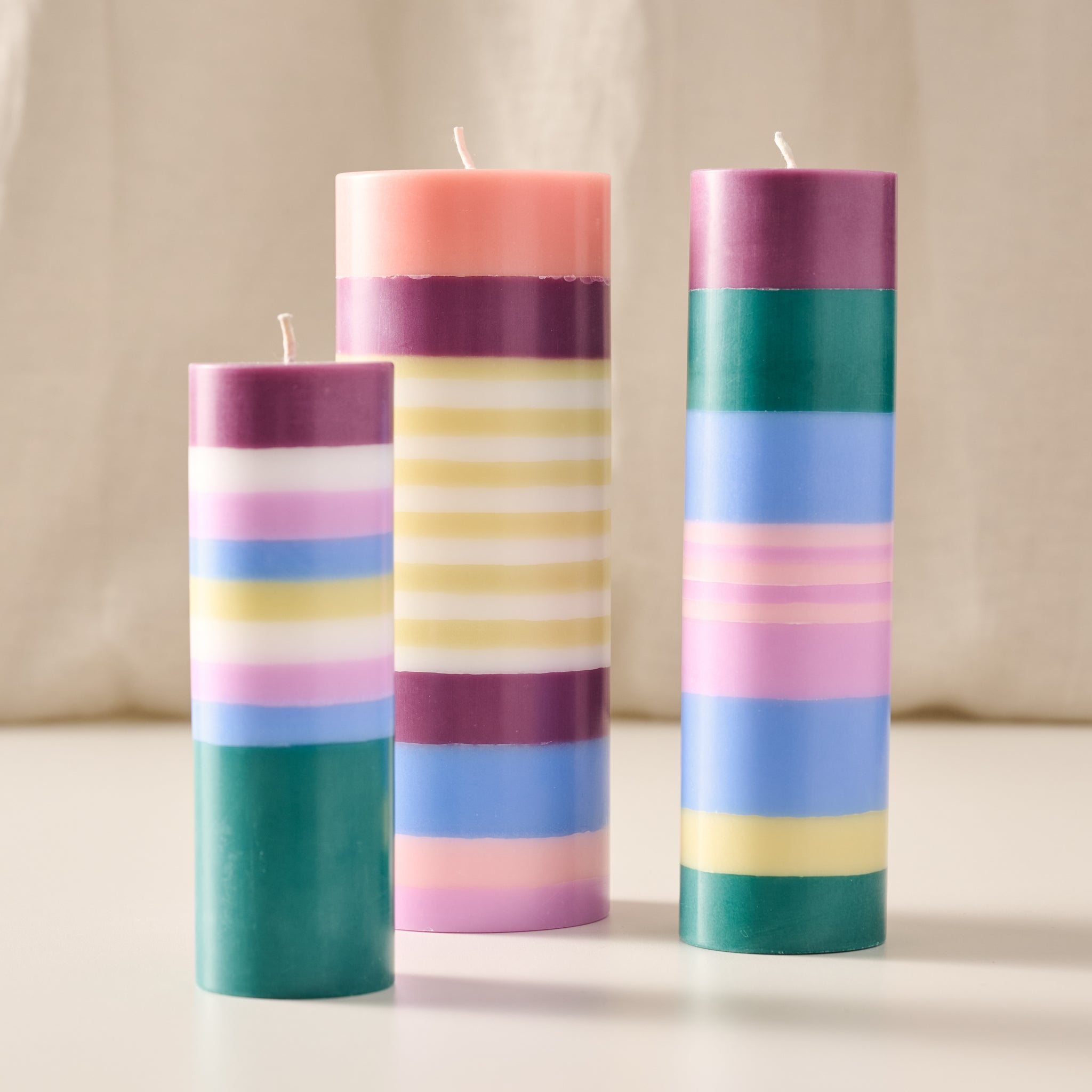 Three striped pillar candles in pastel colors, including pink, purple, teal, blue, yellow, and white, standing on a light surface against a soft beige fabric backdrop.
