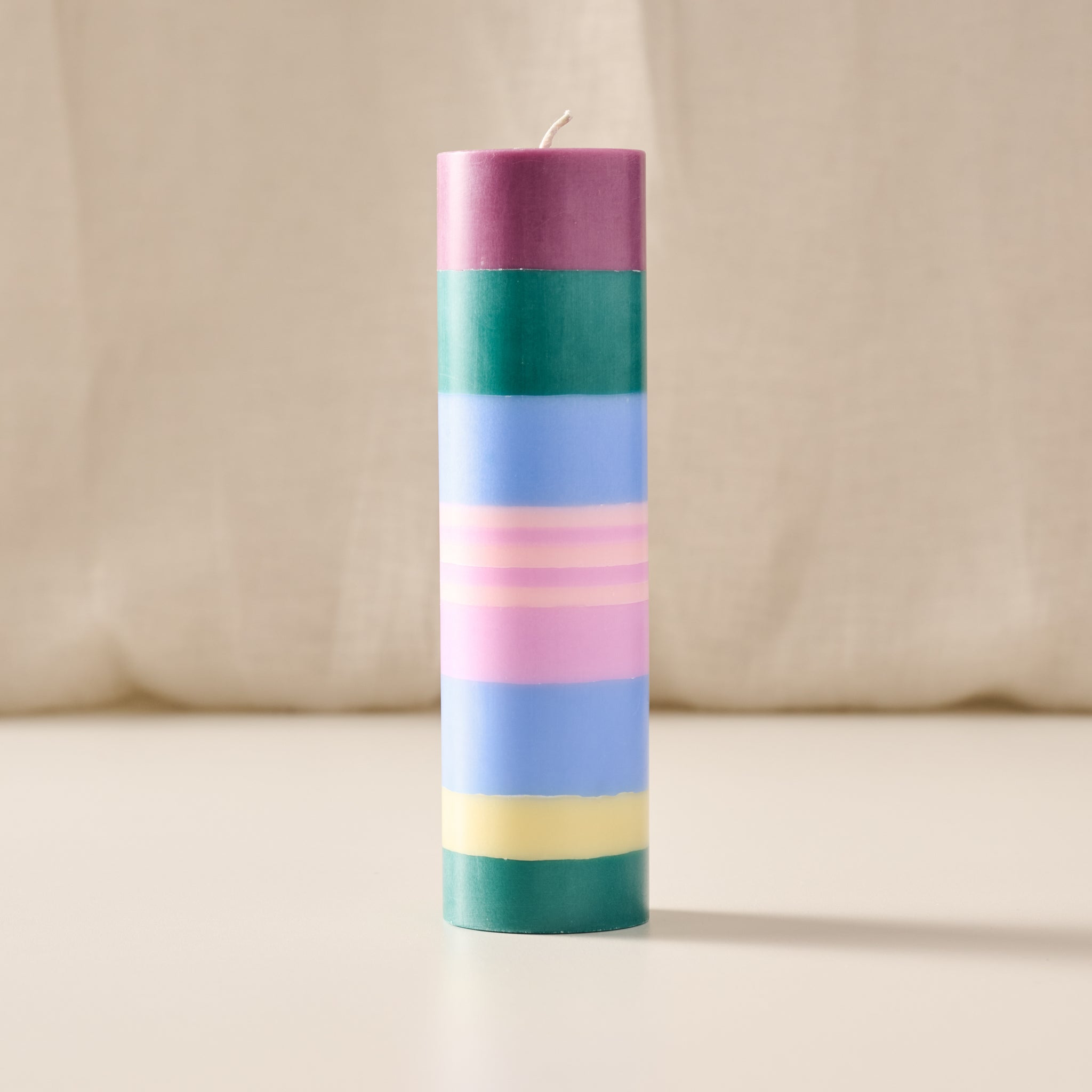 A tall cylindrical candle with horizontal pastel stripes in purple, green, blue, pink, and yellow, standing upright on a light surface against a soft beige fabric background.
