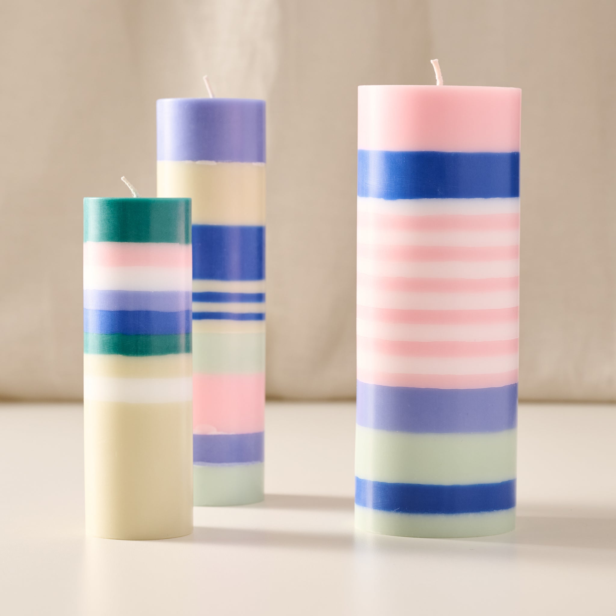 Three striped taper candles in pastel colors, including pink, blue, purple, green, and cream, arranged on a light surface against a soft neutral background.