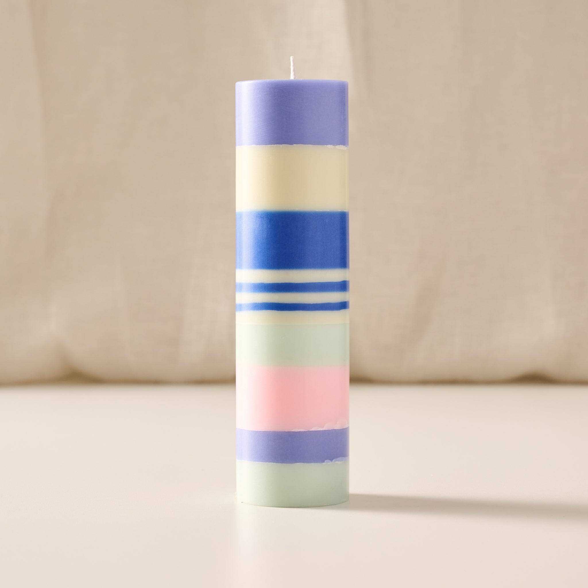A tall, cylindrical striped candle with pastel bands of purple, cream, blue, white, mint green, and pink, standing upright on a light beige surface against a soft cream fabric backdrop.