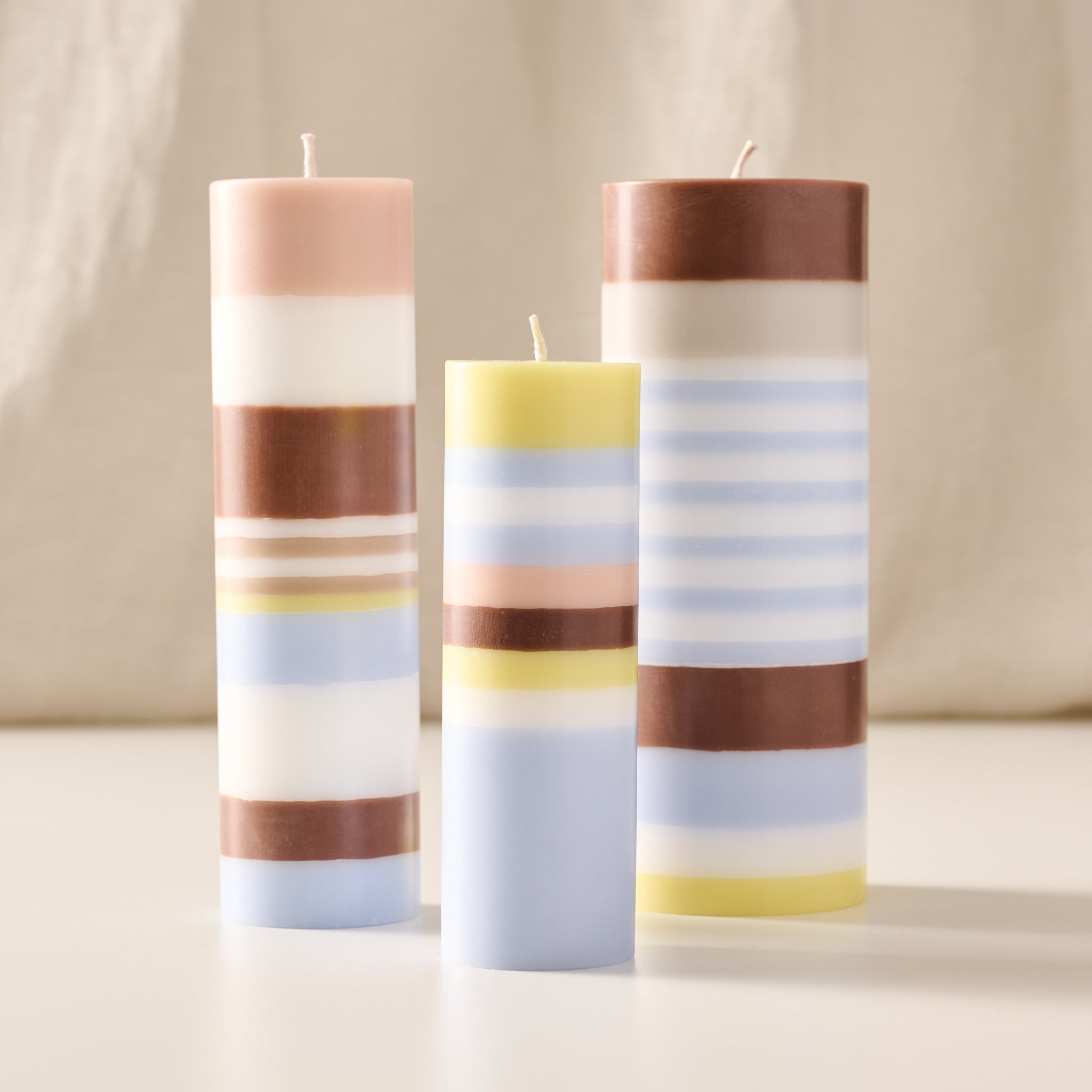 Three striped pillar candles in pastel shades of pink, blue, yellow, and brown, standing on a white surface against a soft beige fabric backdrop.