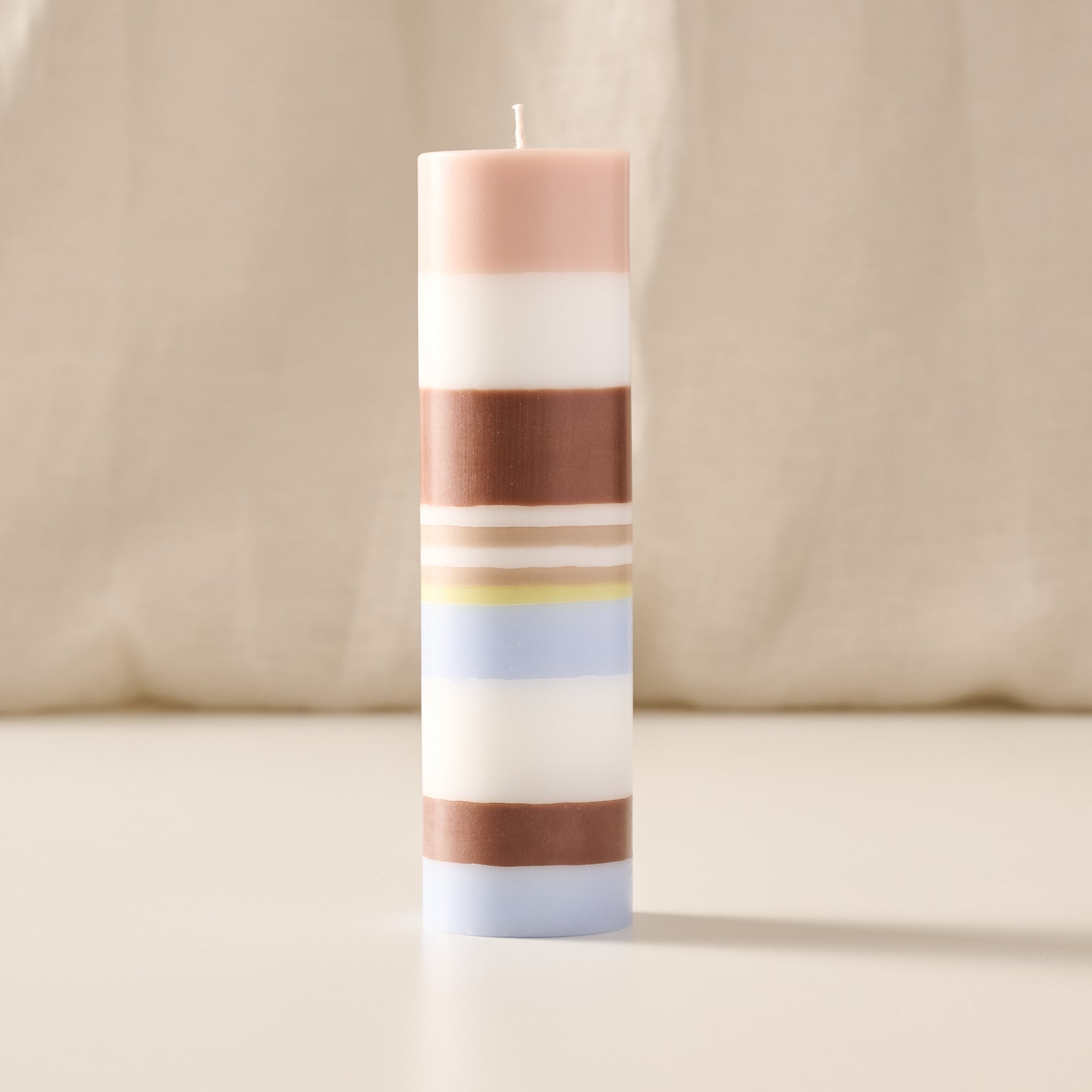 A tall cylindrical candle with horizontal stripes in blush pink, white, brown, pale yellow, light blue, and white, standing upright on a light beige surface against a soft cream fabric backdrop.