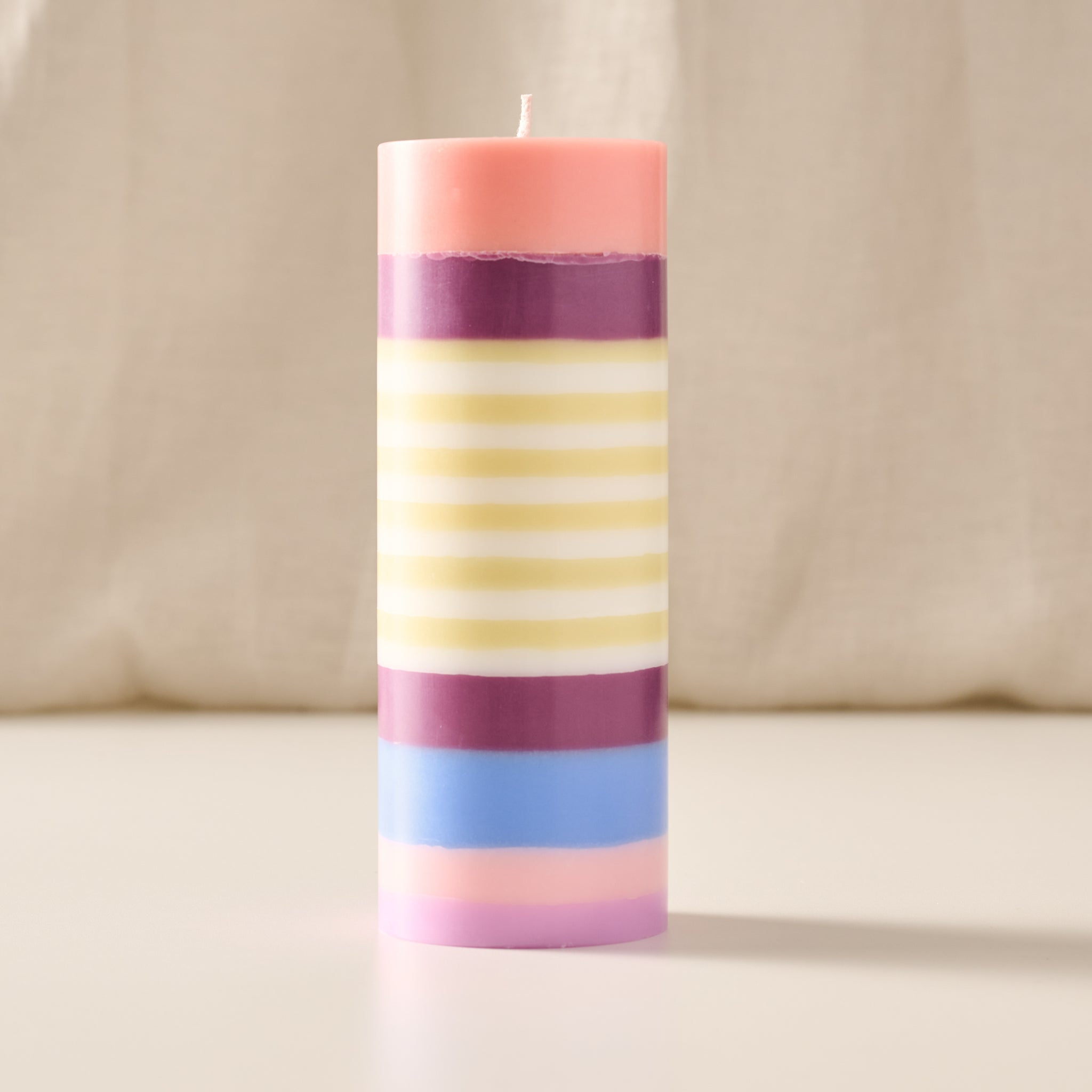 A tall cylindrical candle with horizontal pastel stripes in pink, purple, yellow, white, blue, and light pink, standing upright on a light surface against a soft beige fabric background.