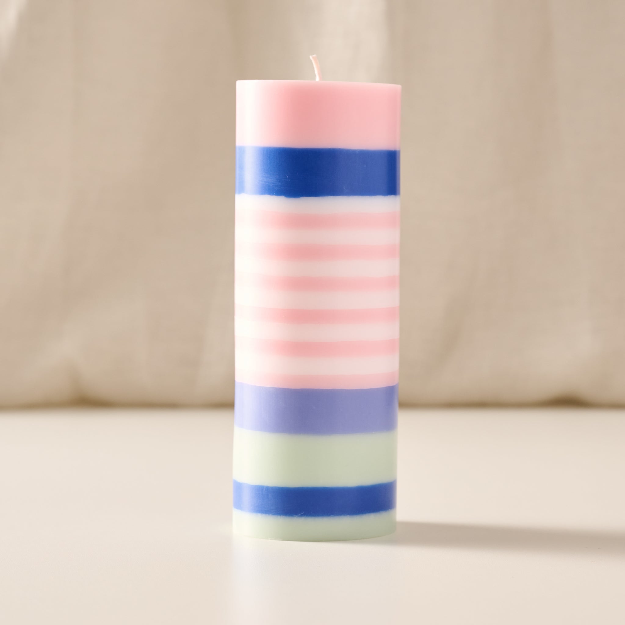 A tall cylindrical pillar candle with horizontal pastel stripes in pink, blue, lavender, and mint green, standing upright on a light surface against a soft beige fabric background.