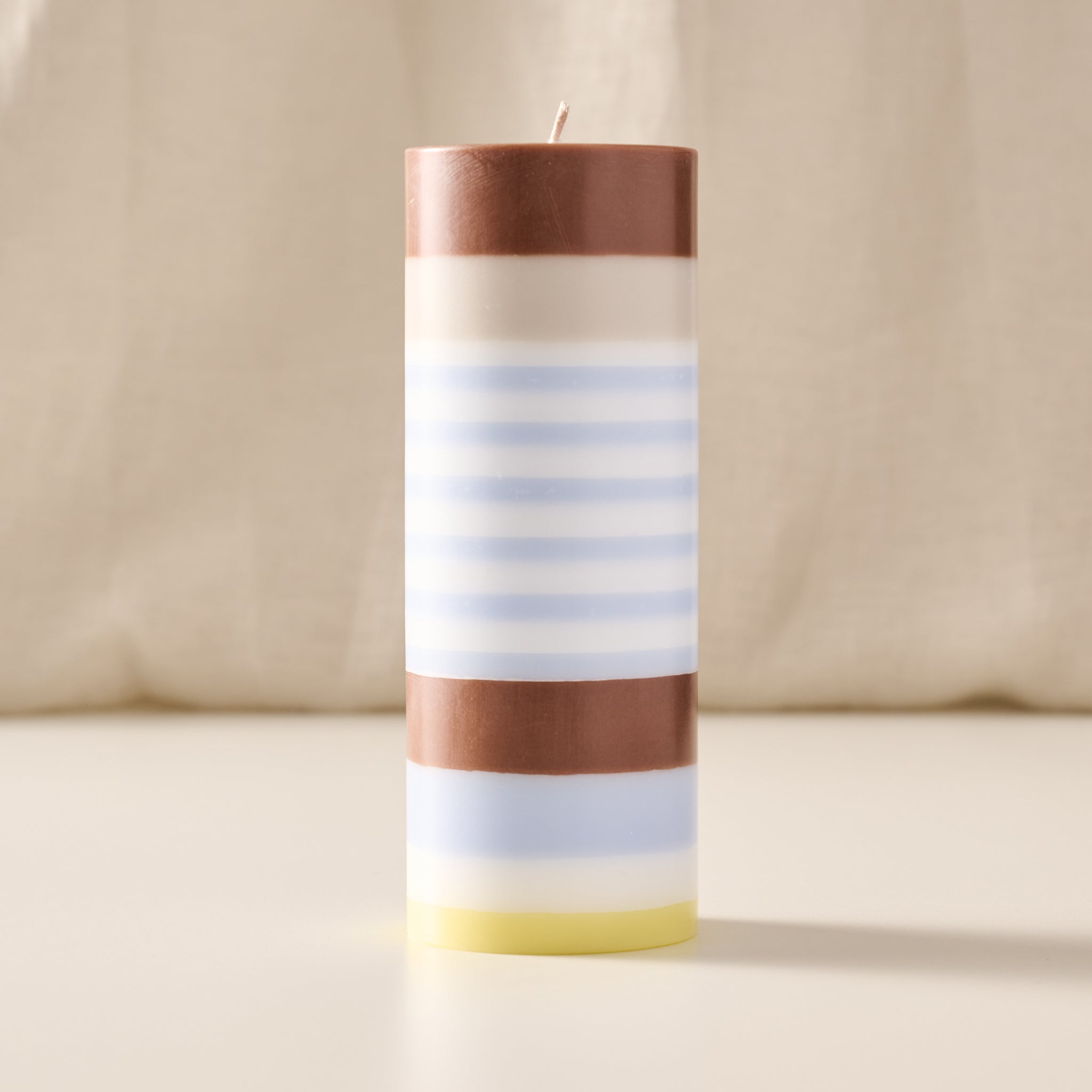 A cylindrical pillar candle with horizontal stripes in brown, cream, light blue, and pale yellow, standing upright on a light surface against a soft beige fabric background.