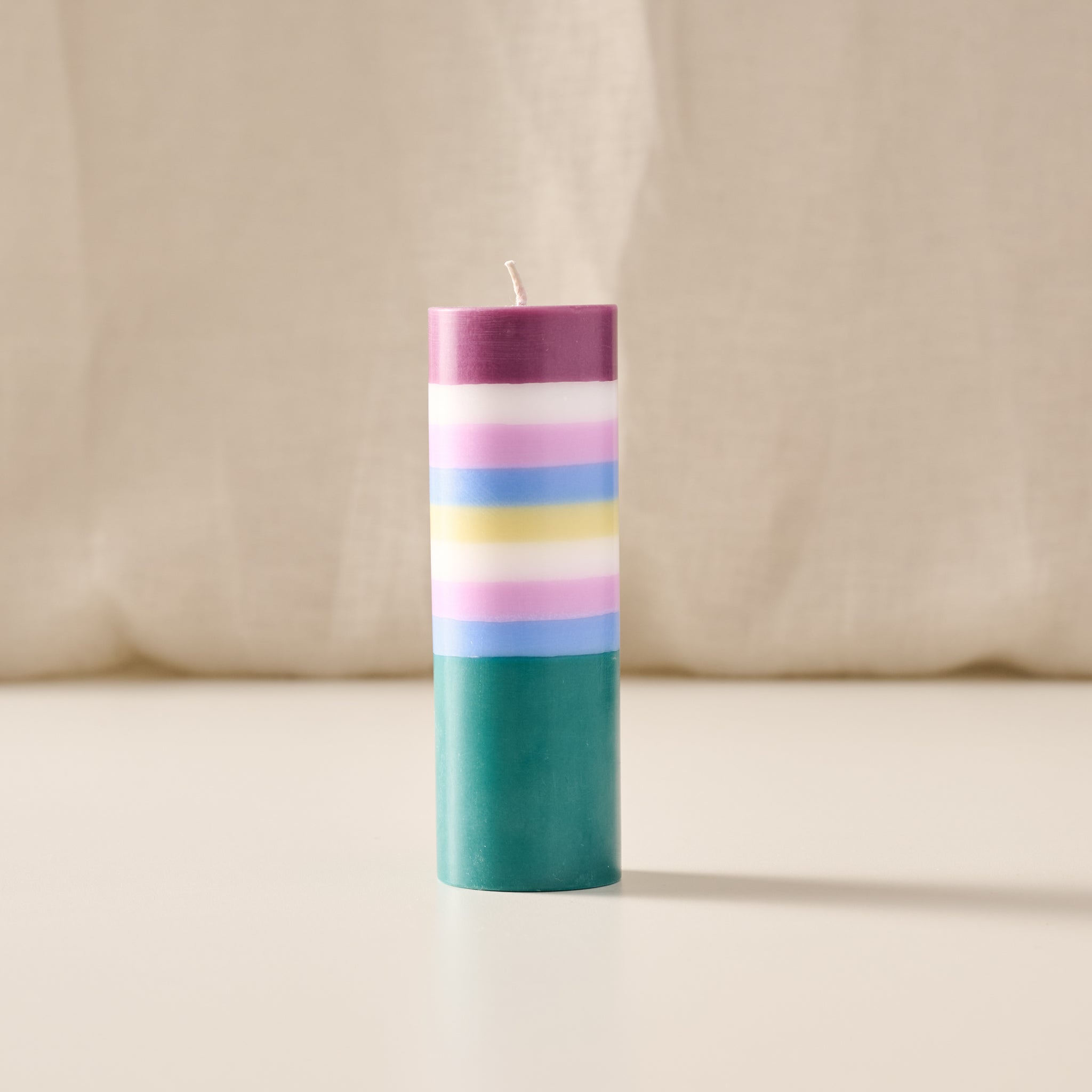 A cylindrical pillar candle with horizontal pastel stripes in purple, white, pink, light blue, yellow, and teal, standing upright on a light beige surface against a soft beige fabric backdrop.