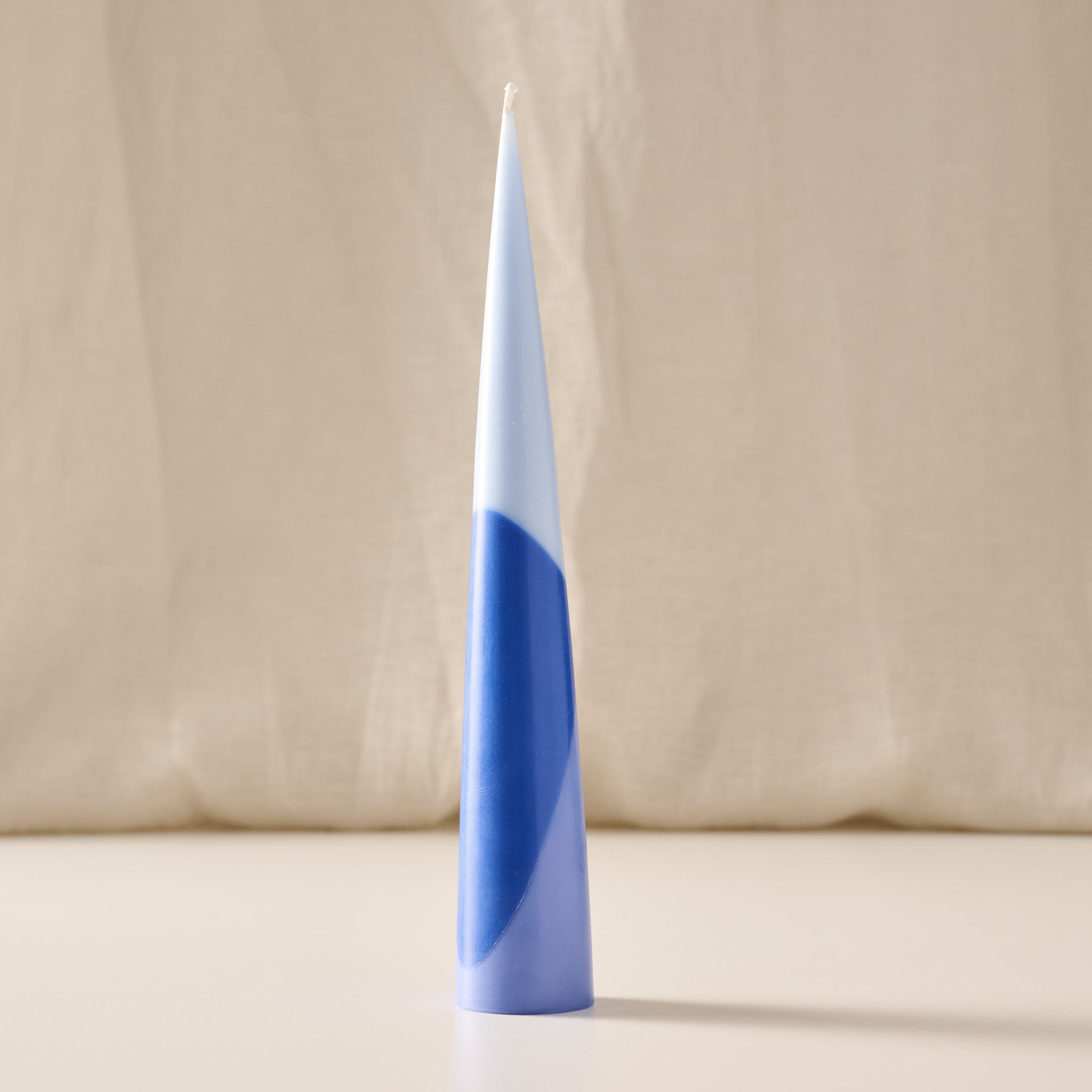 A single taper candle with a modern abstract design, featuring a white upper section and a blue lower section with a wavy pattern, standing upright on a light surface against a draped beige fabric background.