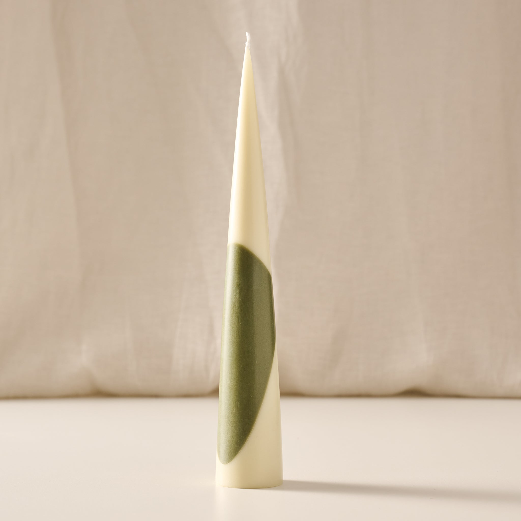 A tall, tapered cream-colored candle stands upright on a light surface against a soft beige fabric backdrop. The candle features an abstract olive-green curved shape wrapping around its lower half. Warm, diffused lighting creates a minimalist, sculptural appearance with a subtle shadow cast to the right.