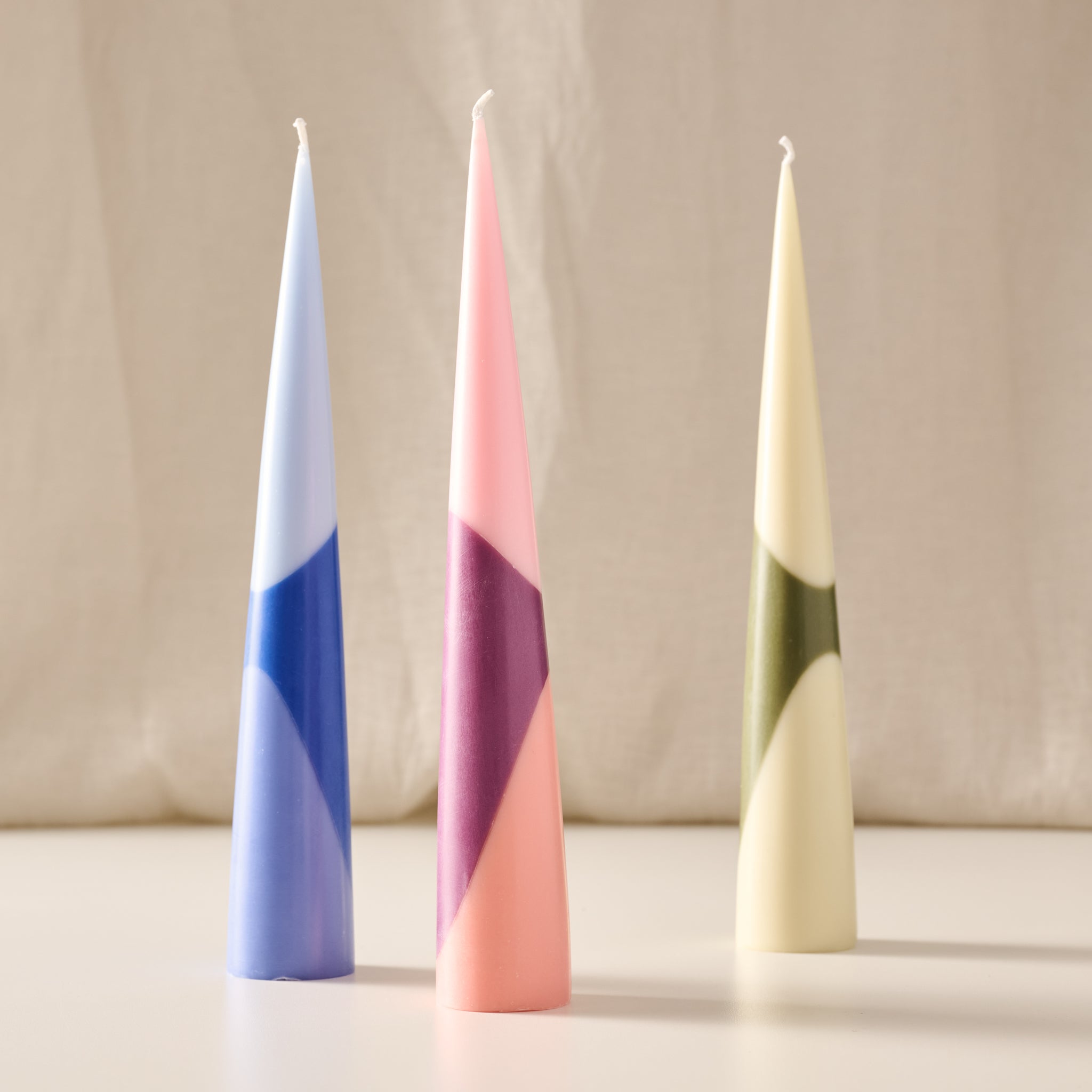Three tapered decorative candles with geometric color-blocked patterns, standing upright on a light surface against a soft beige fabric backdrop. The candles are light blue and navy, pink with purple and peach sections, and cream with olive green sections.