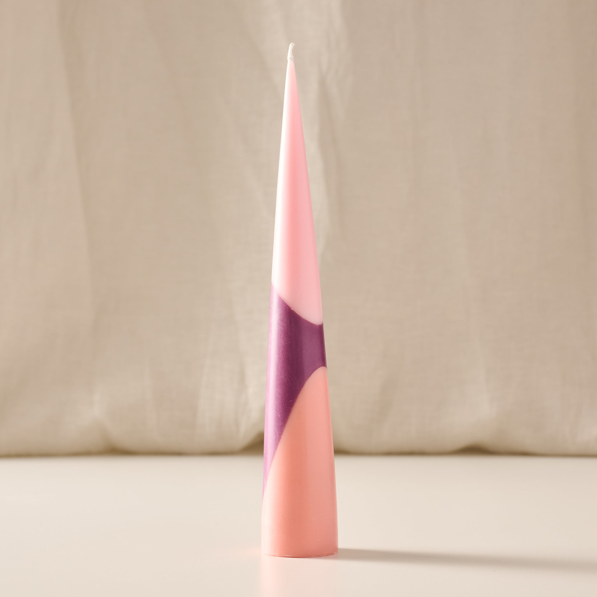 A tall, slender cone-shaped candle with a geometric color-blocked design in shades of pink, peach, and purple, standing upright on a light surface against a soft beige fabric backdrop.