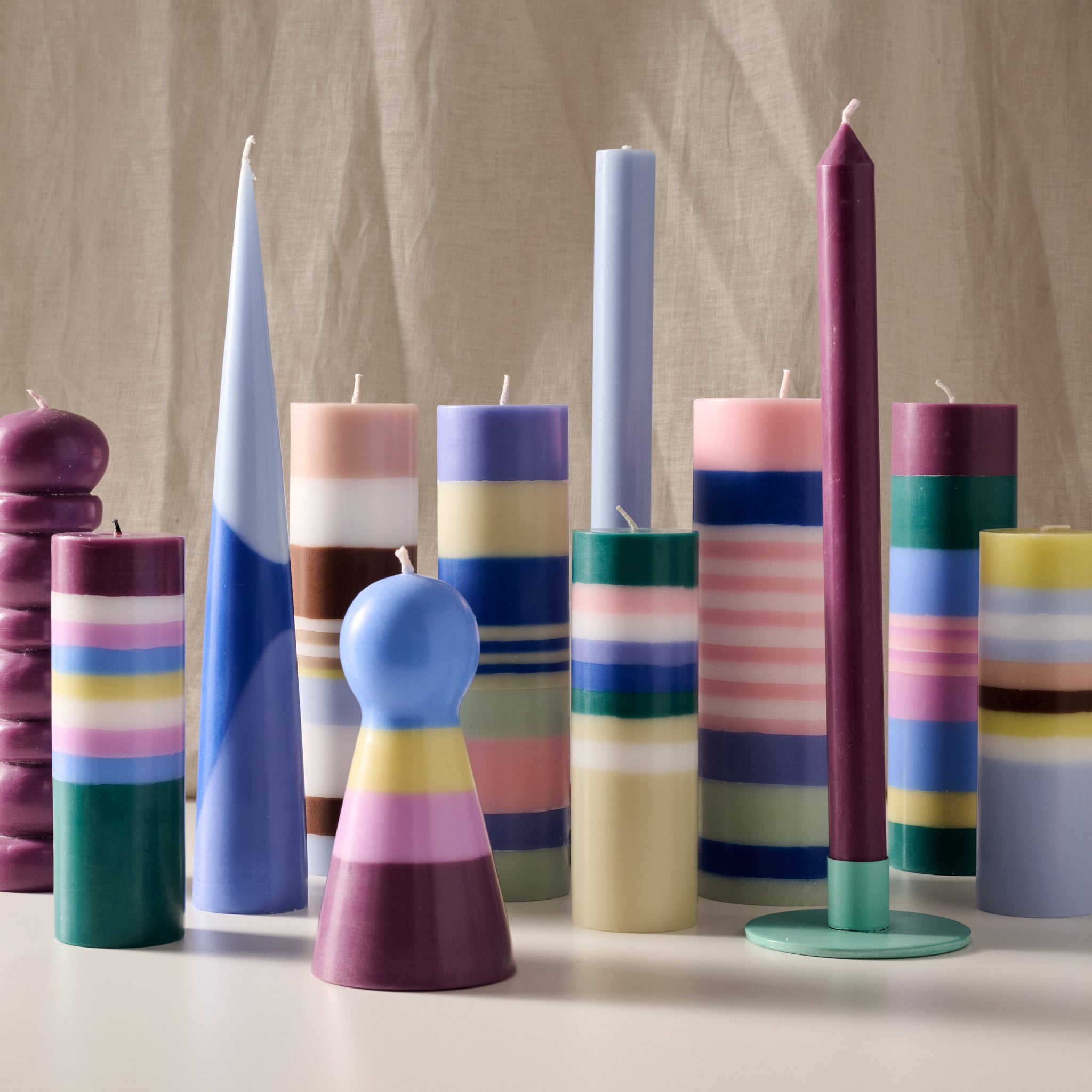 A group of colorful decorative candles and candle holders arranged on a tabletop against a neutral beige fabric backdrop. The set includes striped pillar candles, solid-color candles, and sculptural shapes in pastel and bright hues such as blue, purple, pink, green, yellow, and white.