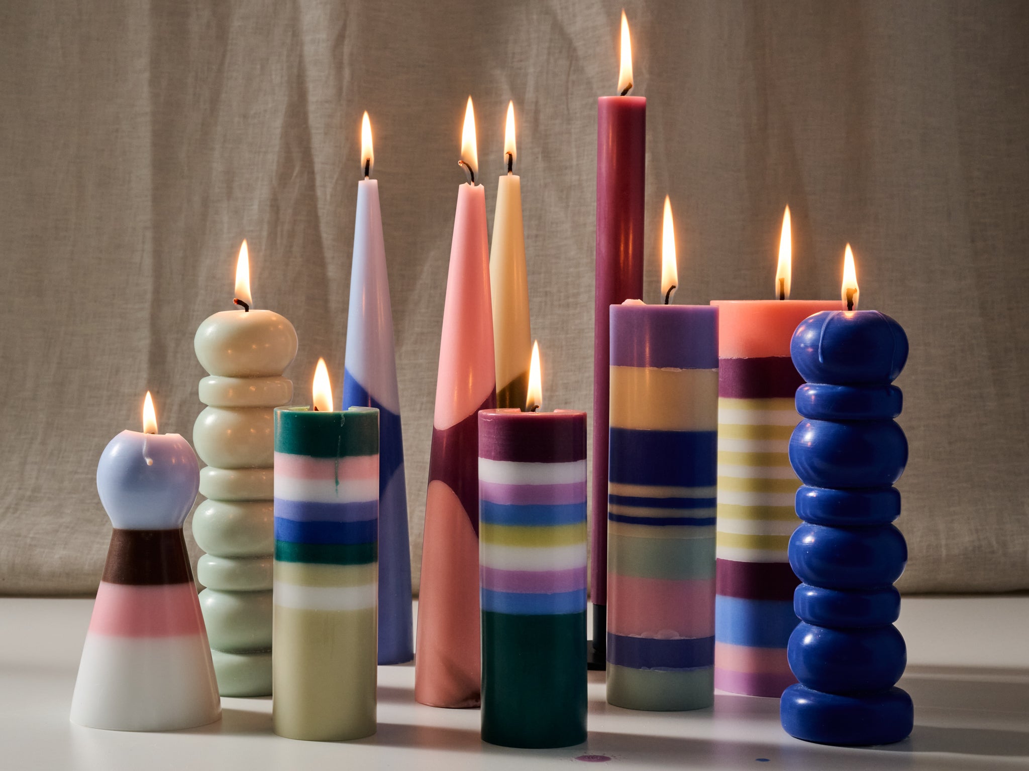 A group of colorful sculptural pillar candles with lit flames, arranged on a white surface against a neutral beige fabric backdrop. The candles feature various geometric and striped designs in shades of blue, pink, green, yellow, and purple.
