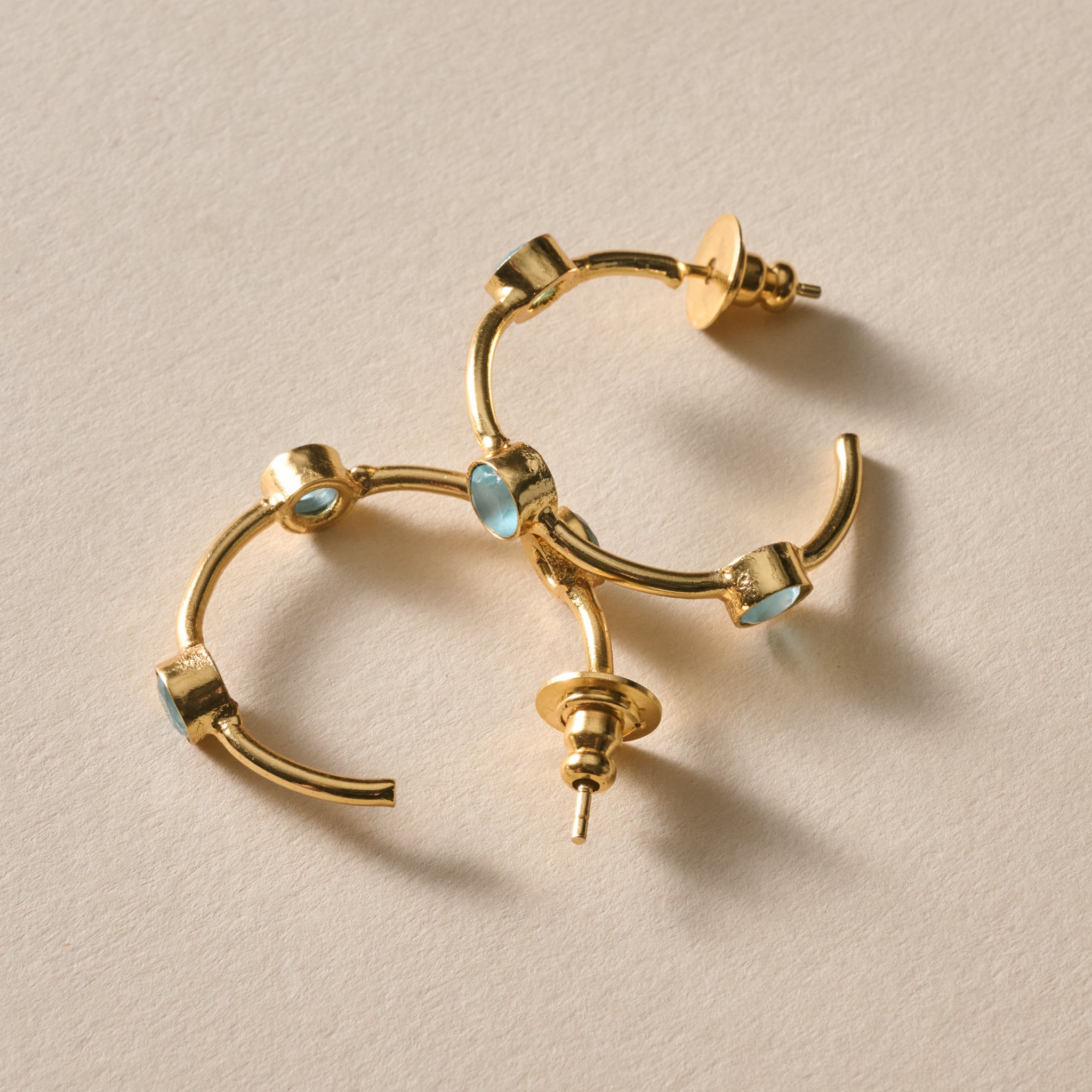 Gold hoop earrings with small light blue oval inlays, photographed on a plain light beige surface.