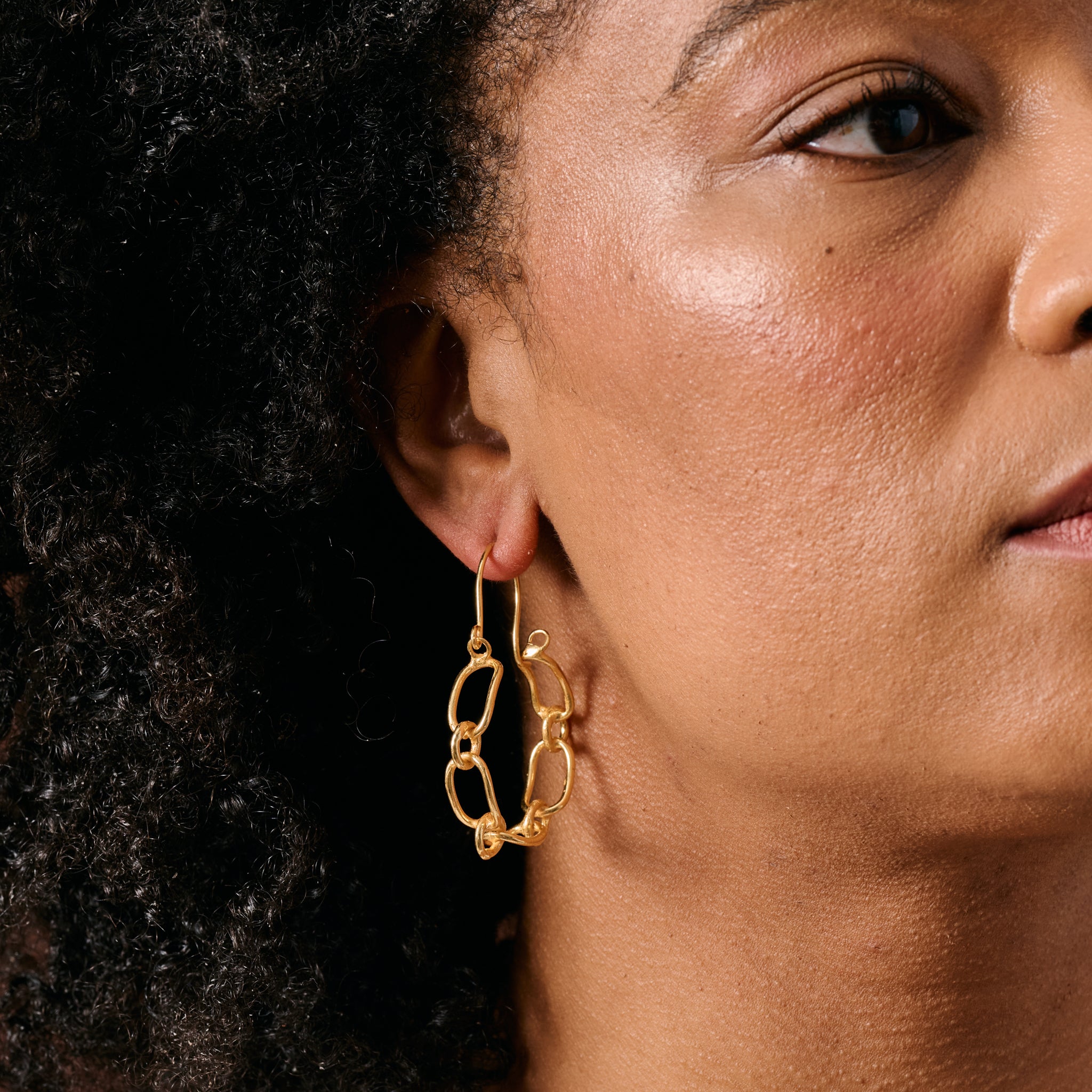 A close-up of a woman wearing a gold hoop earring with an open, chain-link style design.