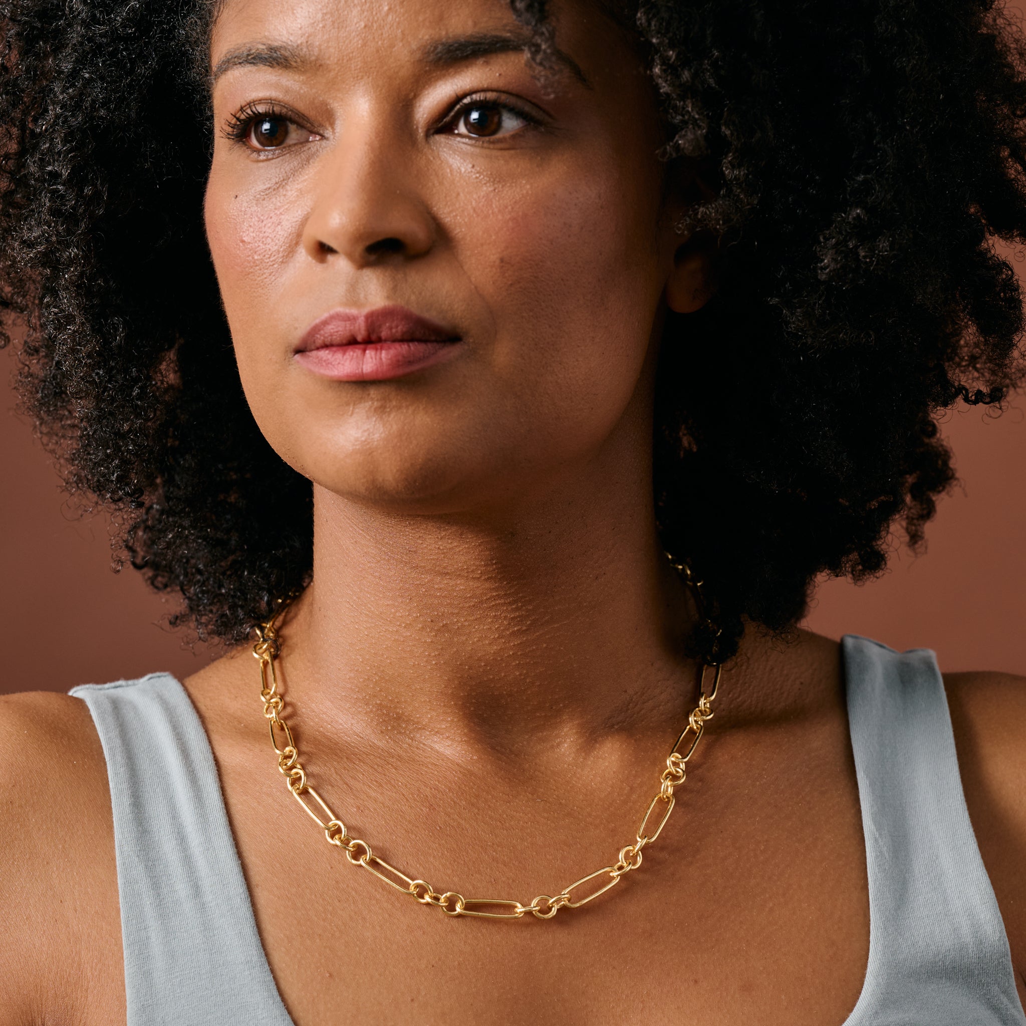 A close-up of a woman wearing a gold chain link necklace and a light blue top, set against a solid warm brown background.