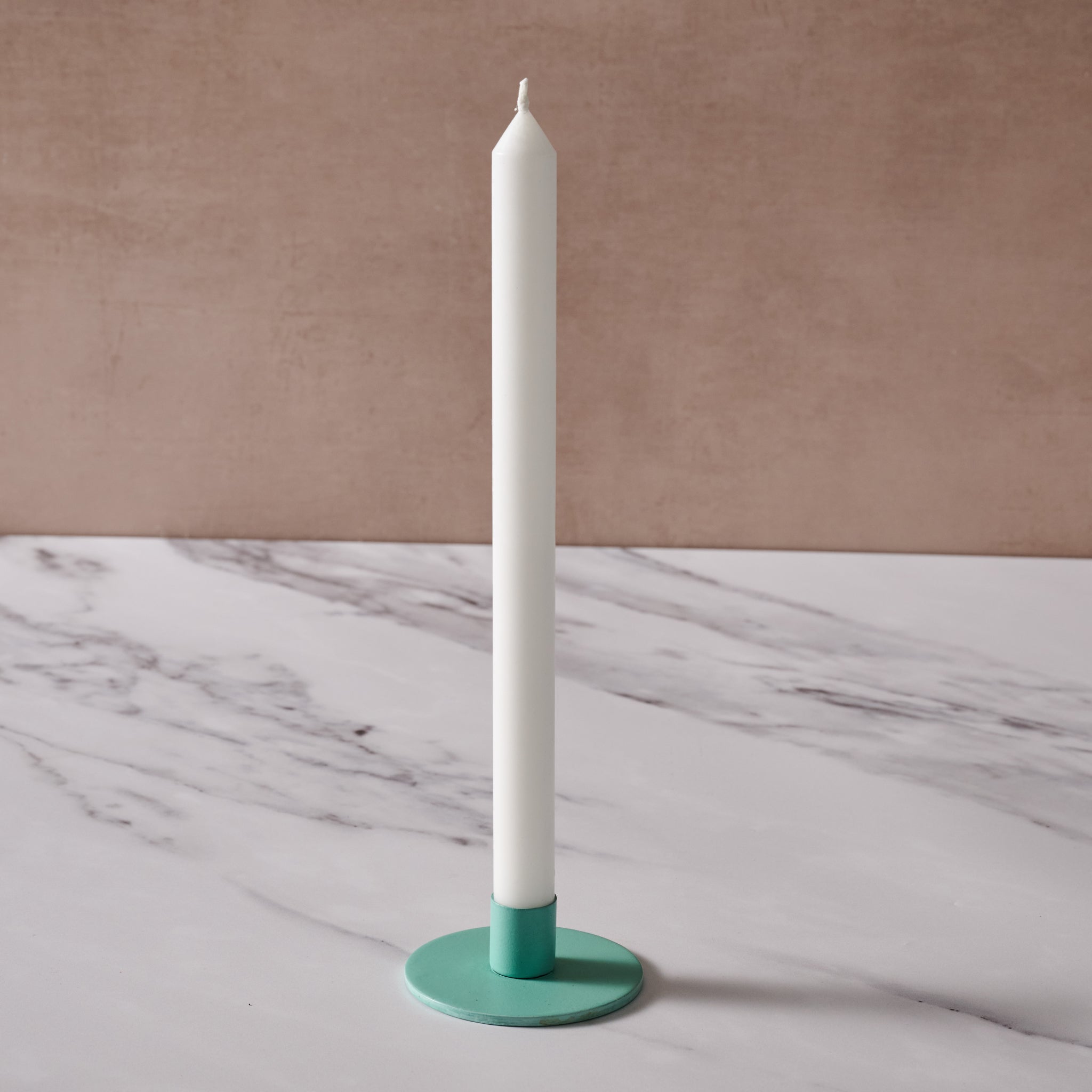 A white taper candle in a round turquoise holder, set on a white marble surface with a textured beige background.