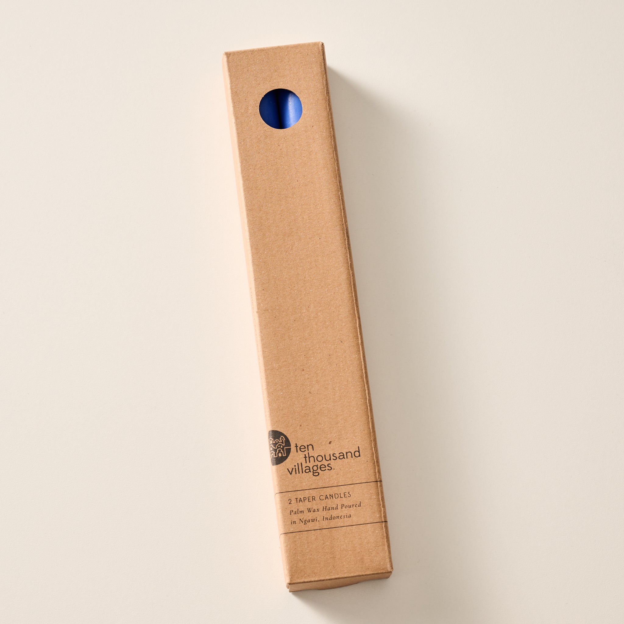 A tall, narrow cardboard box with a circular window display cutout at the top showing two cobalt blue candles inside, photographed on a clean off-white background. The box has printed text near the bottom including the brand name 'Ten Thousand Villages'.