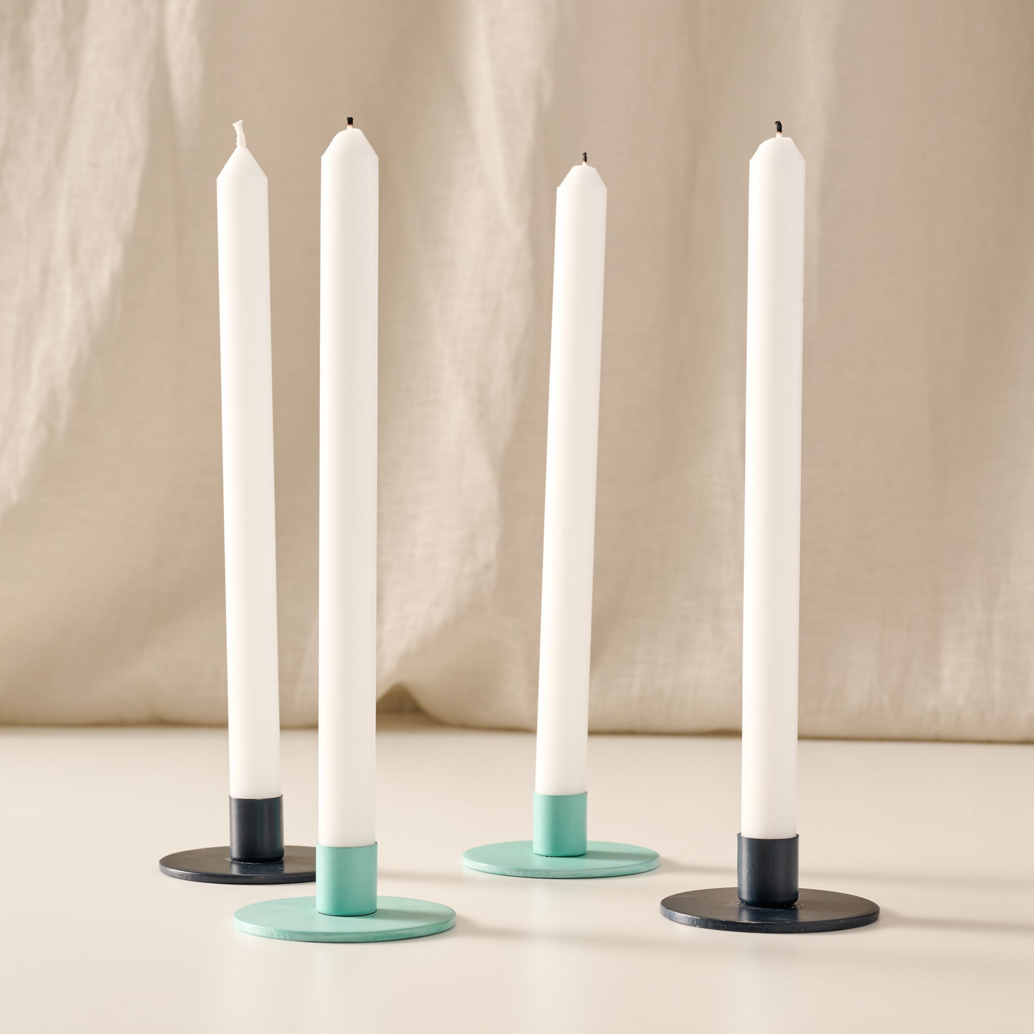 Four taper candles in white holders with round turquoise and blue bases, arranged on a light surface against a soft beige fabric backdrop.