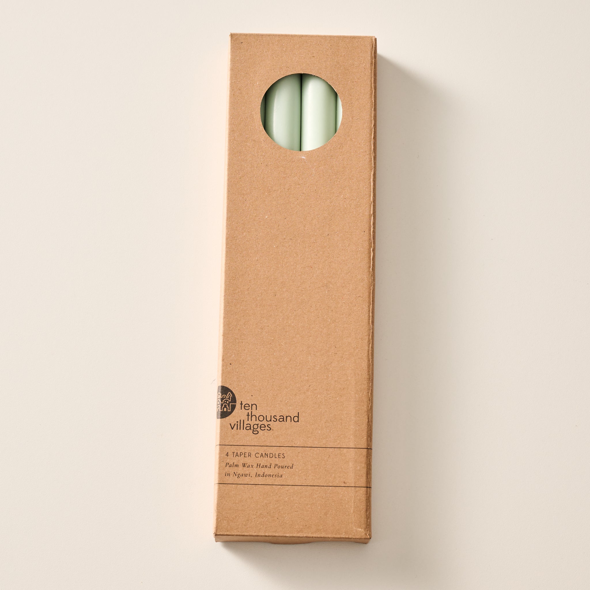 A tall, narrow cardboard box with a circular window display cutout at the top showing two sage green candles inside, photographed on a clean off-white background. The box has printed text near the bottom including the brand name 'Ten Thousand Villages'.