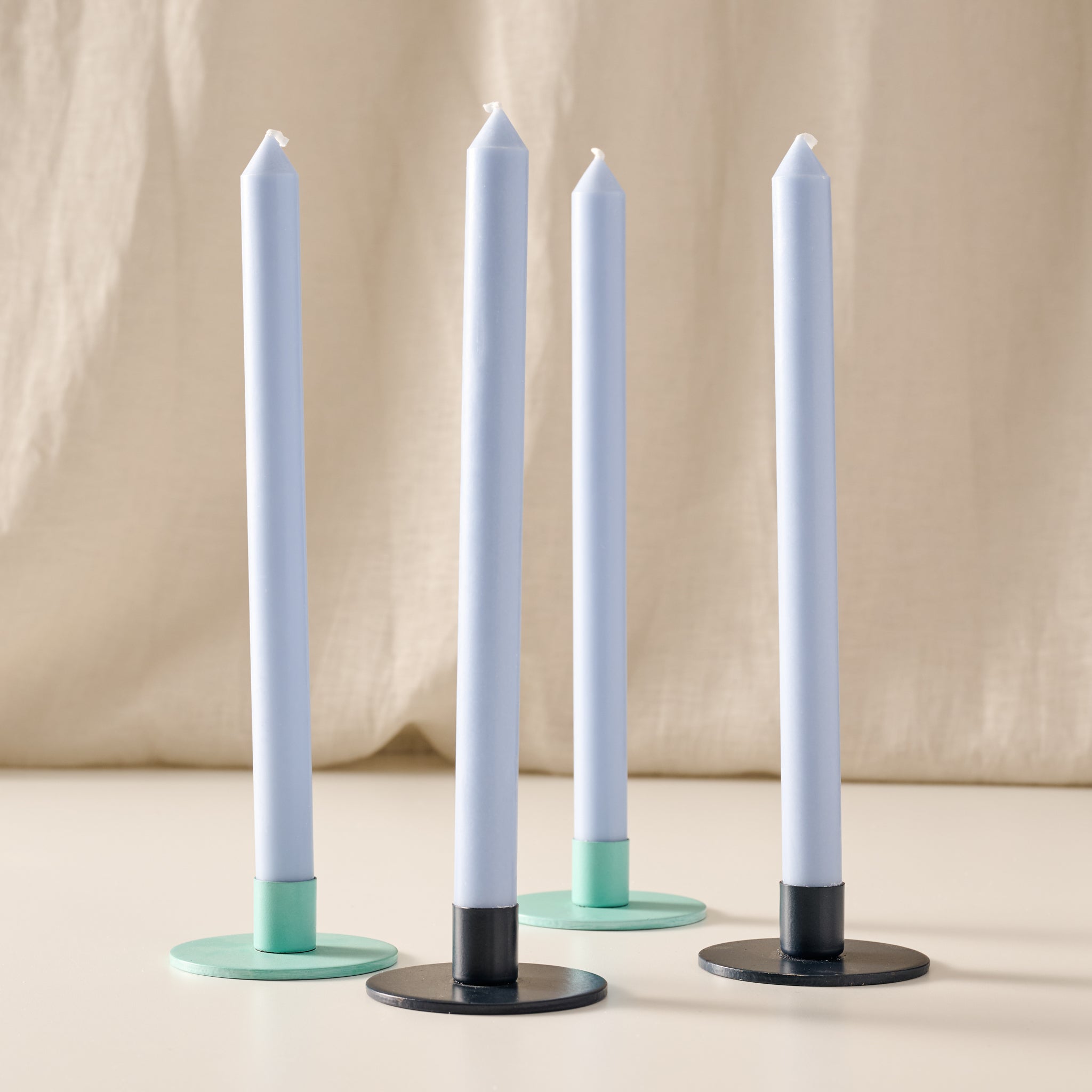 Four taper candles in mint with round metal candle holders in turquoise and blue, arranged on a light beige surface against a draped beige fabric background.
