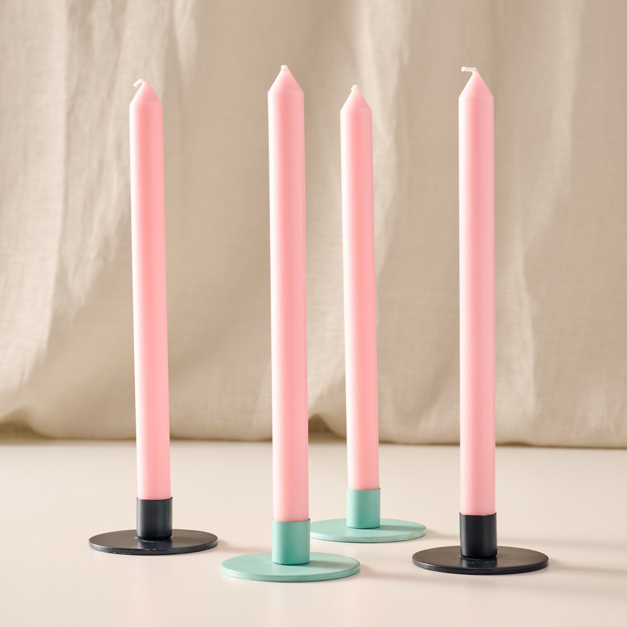 Four taper candles in a ginger color with turquoise and blue candle holder bases, arranged on a light surface against a soft beige fabric backdrop.