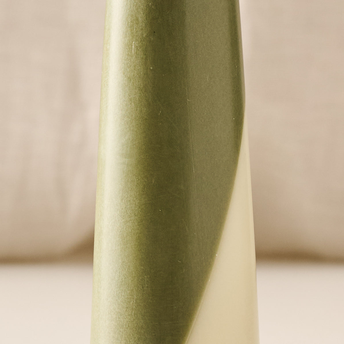 A close-up of a tapered candle with a vertical geometric design in olive green and cream, set against a soft beige background.