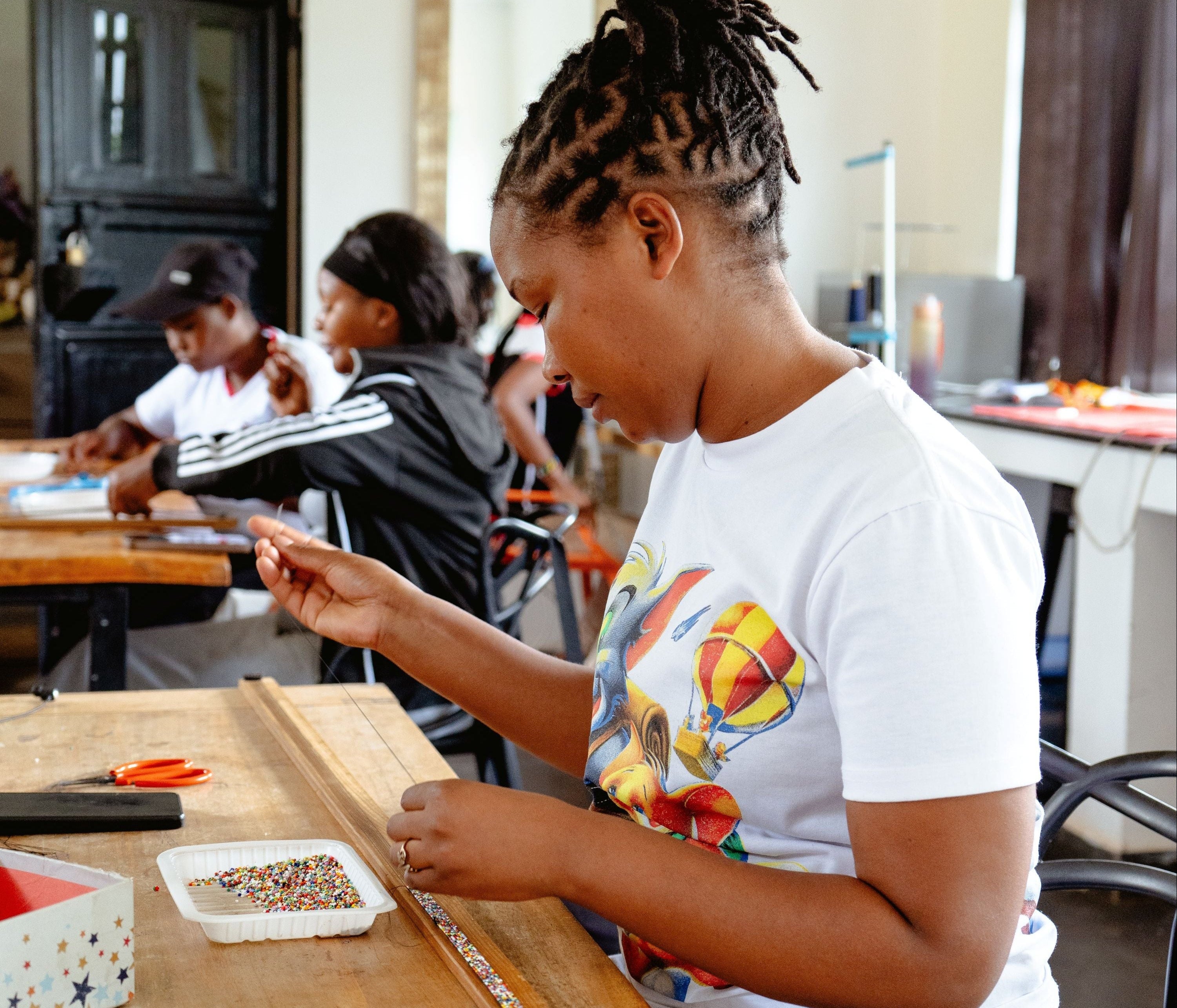 A woman with braided hair sits at a wooden table in a workshop, wearing a white graphic t-shirt while working with craft supplies, including a small container of colorful beads.